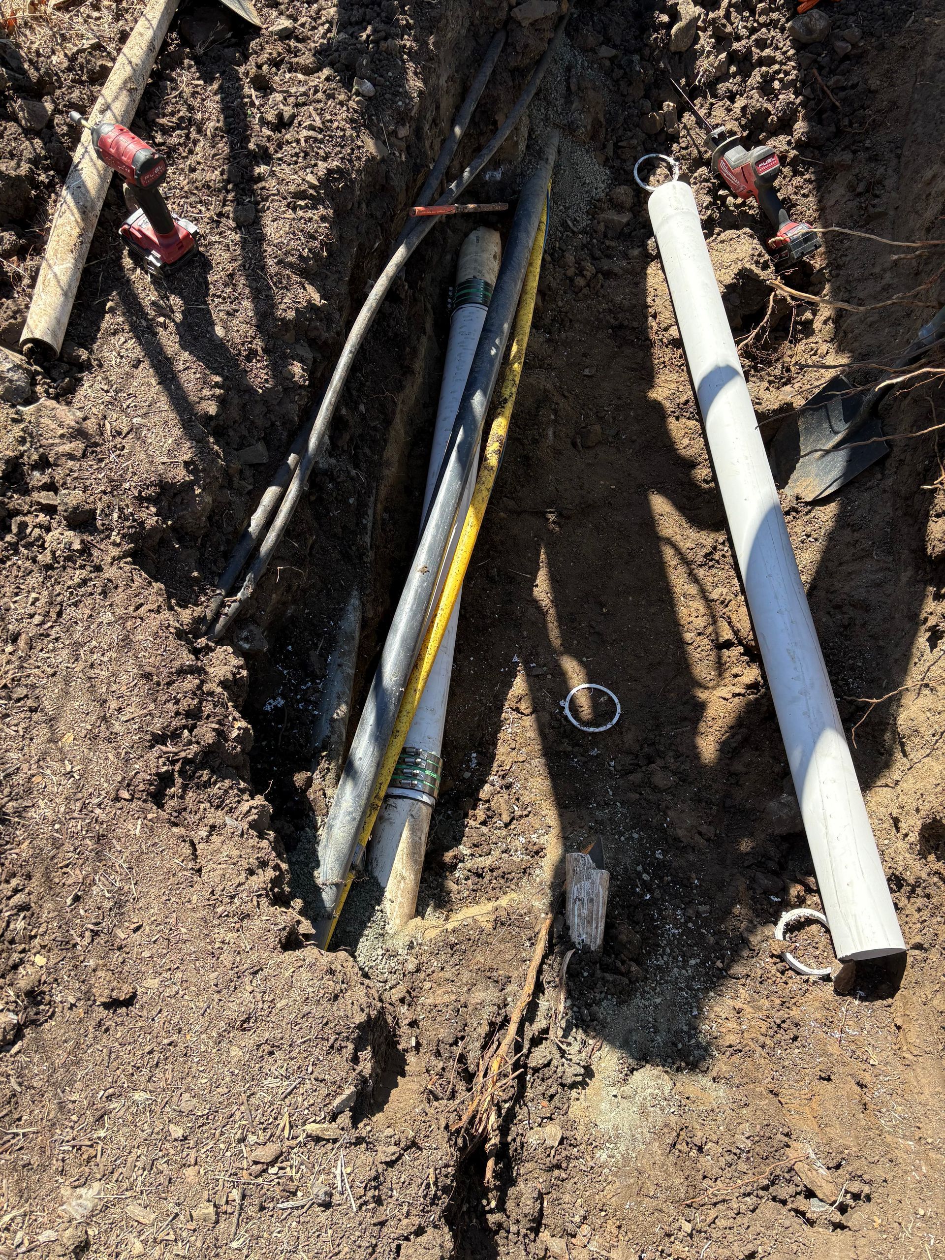 Two grey PVC pipes and a measuring tape lay in a narrow dirt trench during an outdoor construction project.