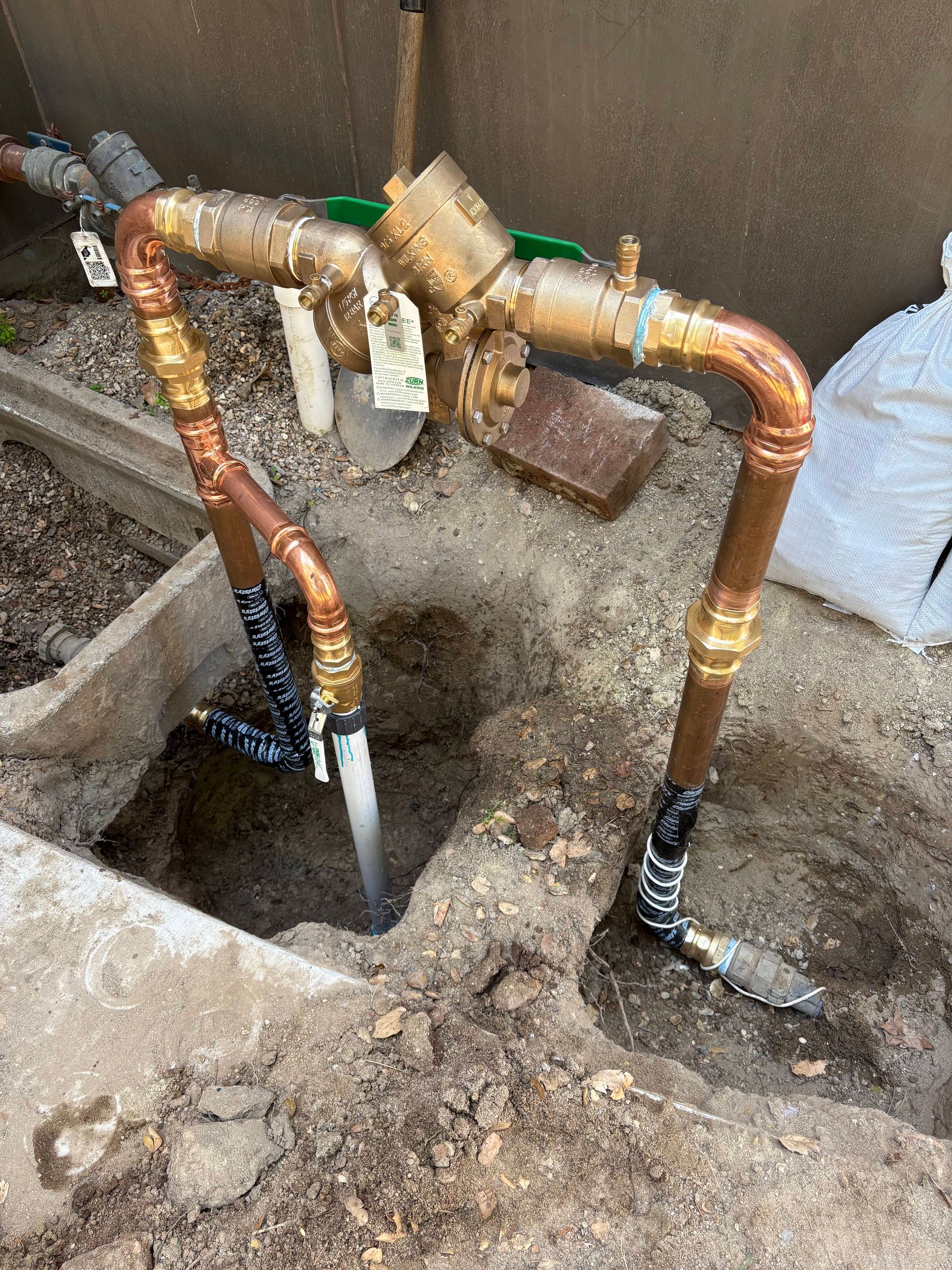 A brass backflow prevention assembly connected to copper pipes, partially installed in a dirt-filled trench.