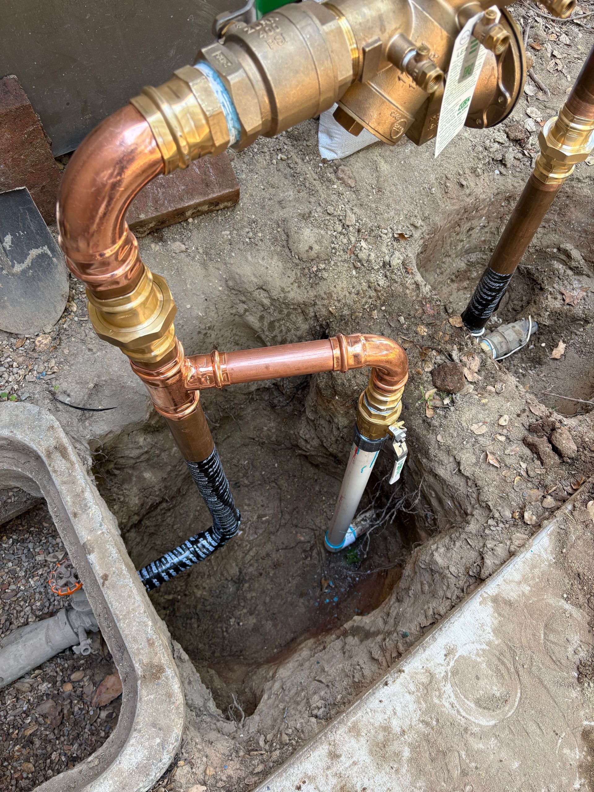Newly installed brass and copper pipe assembly connecting irrigation system components in an outdoor dirt trench.