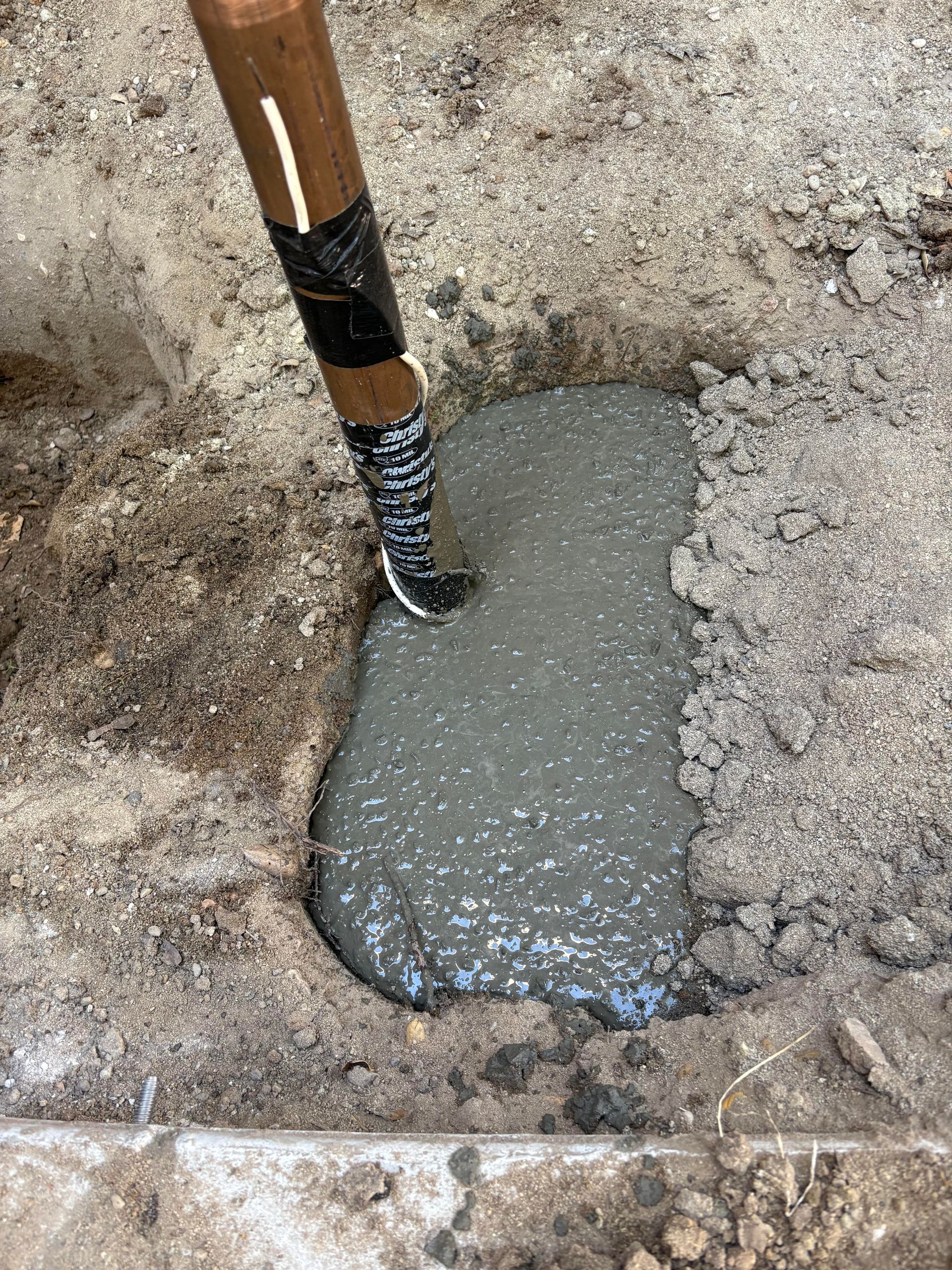 A wooden pole wrapped in black tape is submerged into a hole filled with wet, gray concrete in the ground.