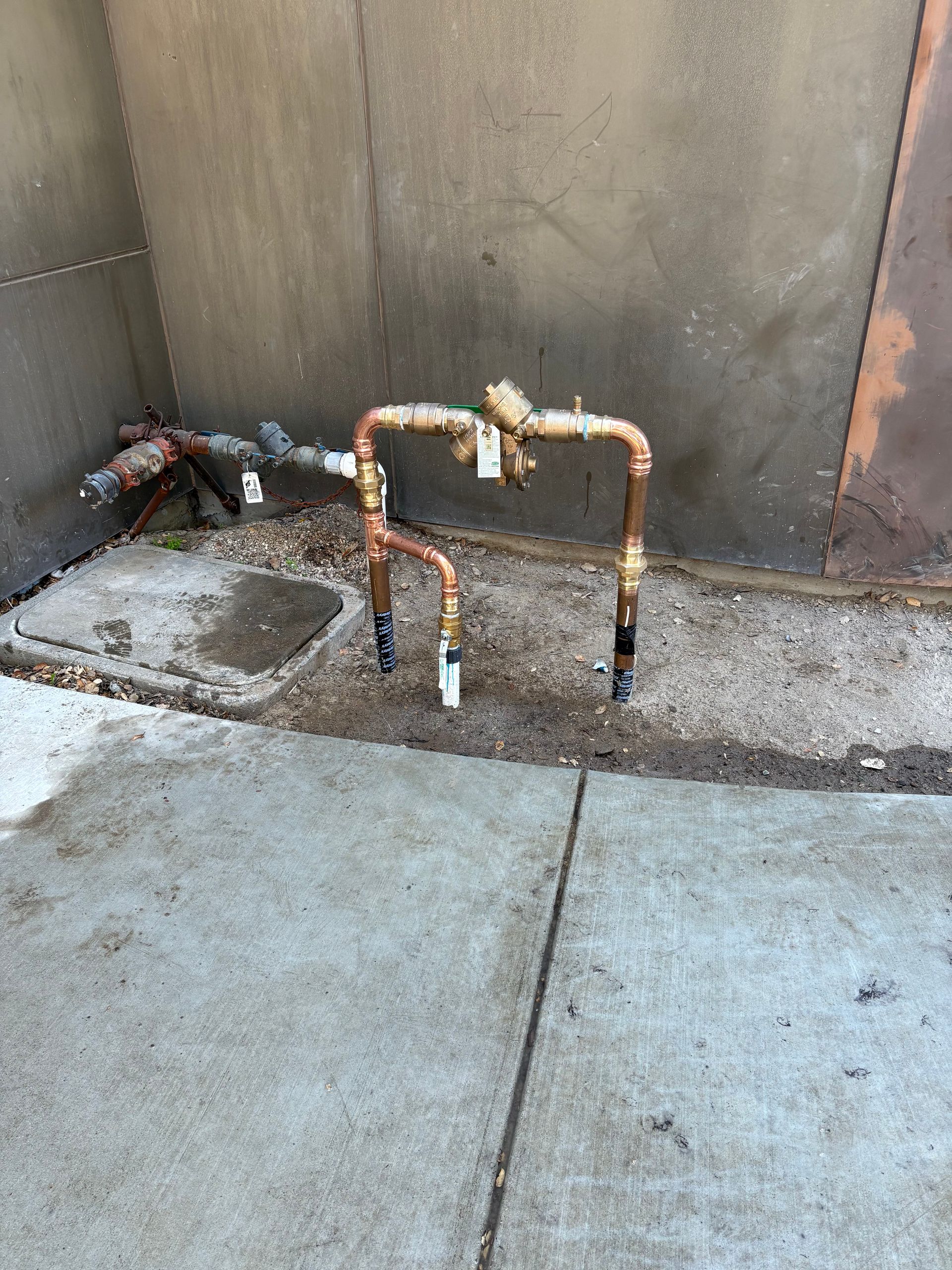 A copper backflow prevention device installed in the ground next to a concrete wall.
