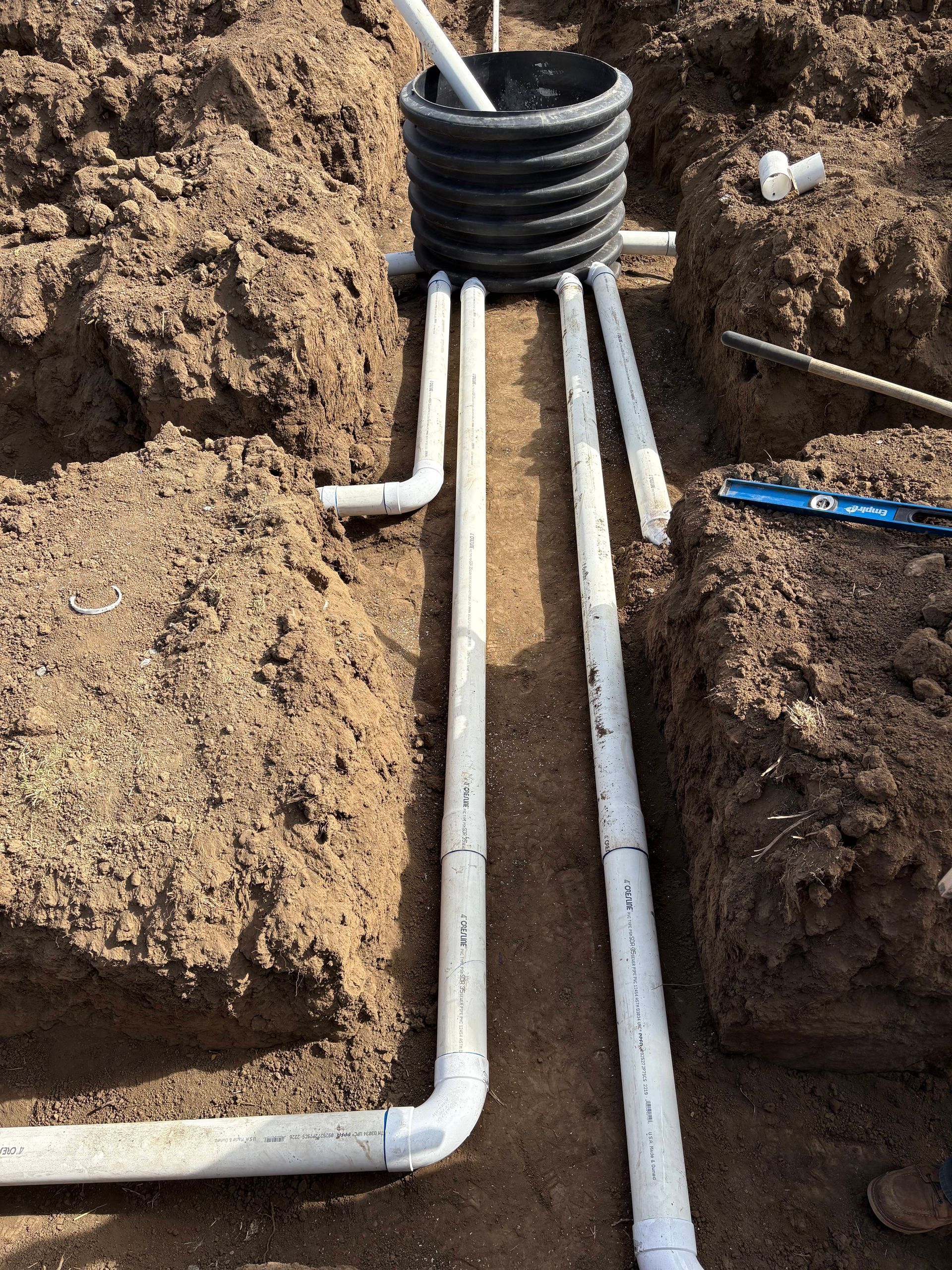 White PVC pipes connect to a black plastic drainage basin in a dirt trench during a construction project.
