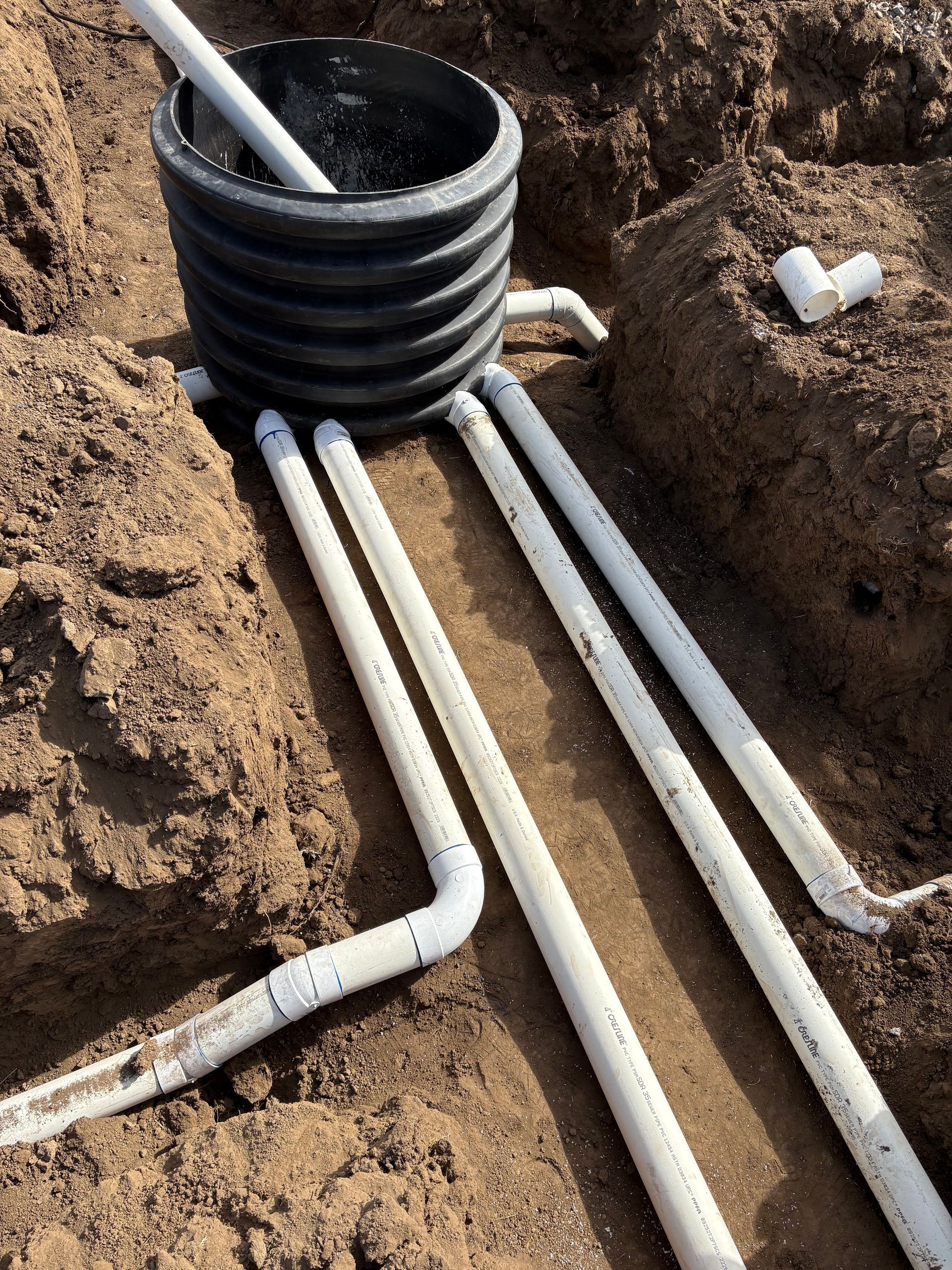 A black corrugated drainage basin connected to multiple white PVC pipes laid in dirt-filled trenches.