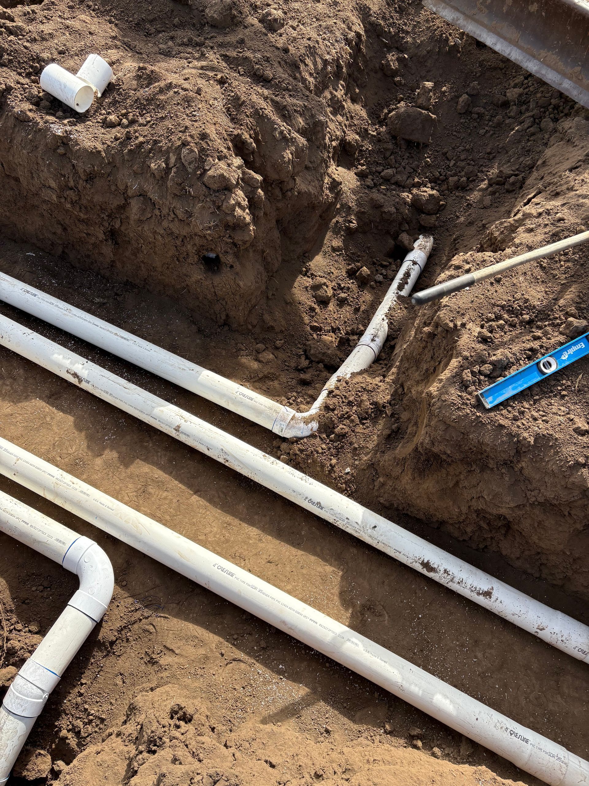 White PVC pipes installed in a narrow, dirt-filled trench during a construction project.