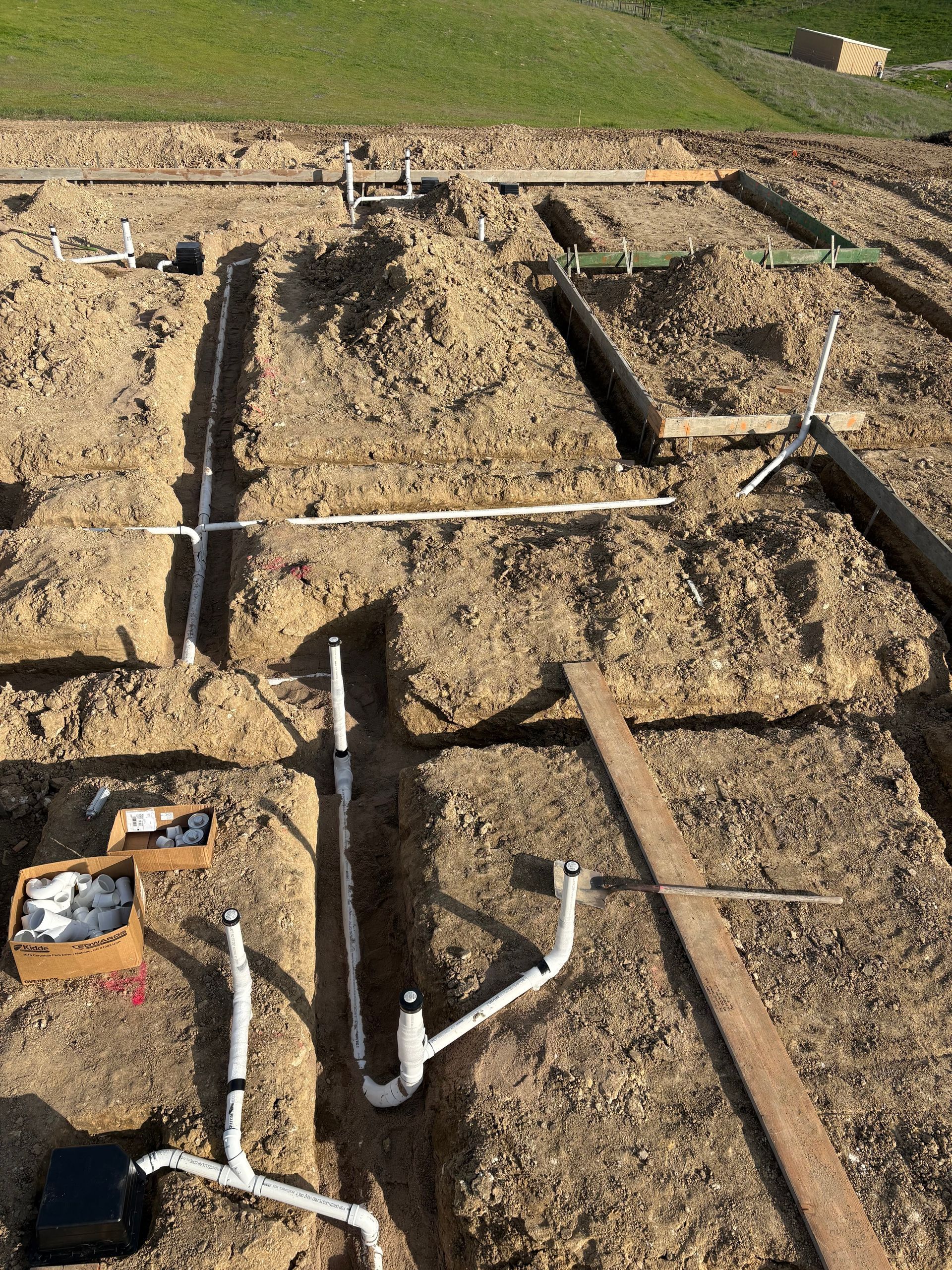 White PVC plumbing pipes laid in narrow dirt trenches for a residential foundation construction site.