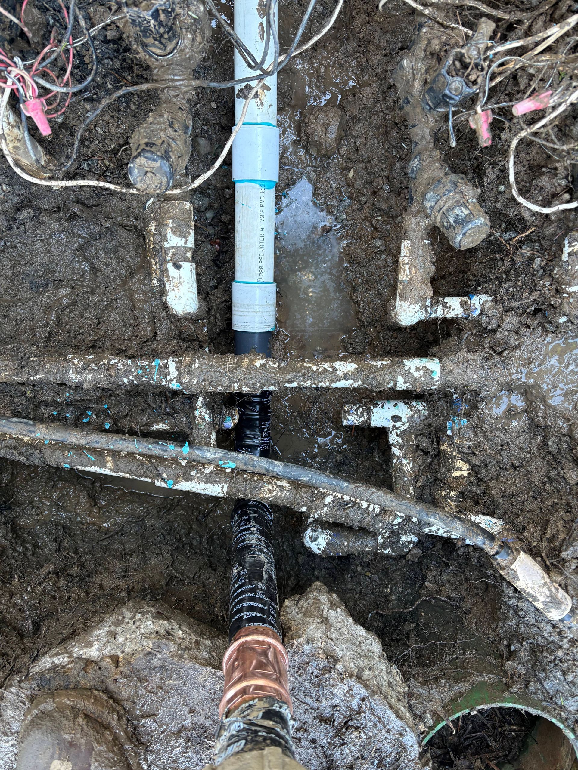 A high-angle view shows an underground repair of irrigation pipes with PVC, black poly tubing, and wiring in wet mud.