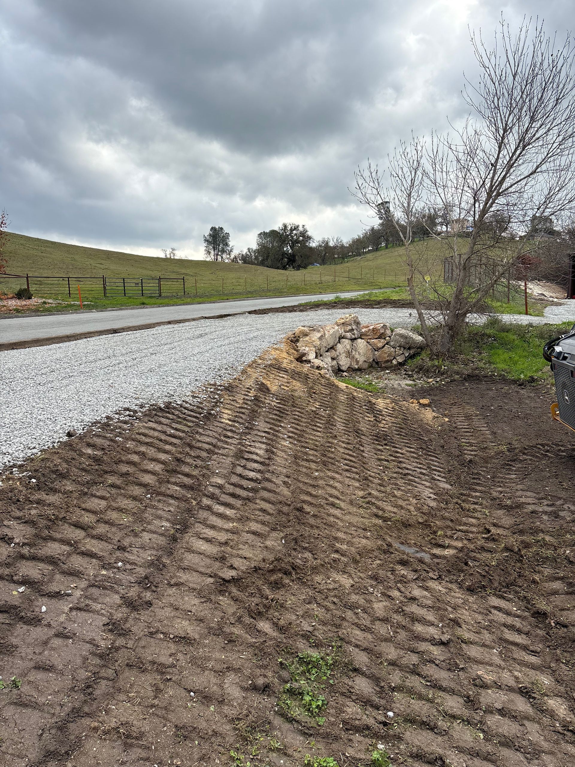A gravel path curves through a grassy field under a cloudy sky, with tire tracks imprinted in the muddy foreground.