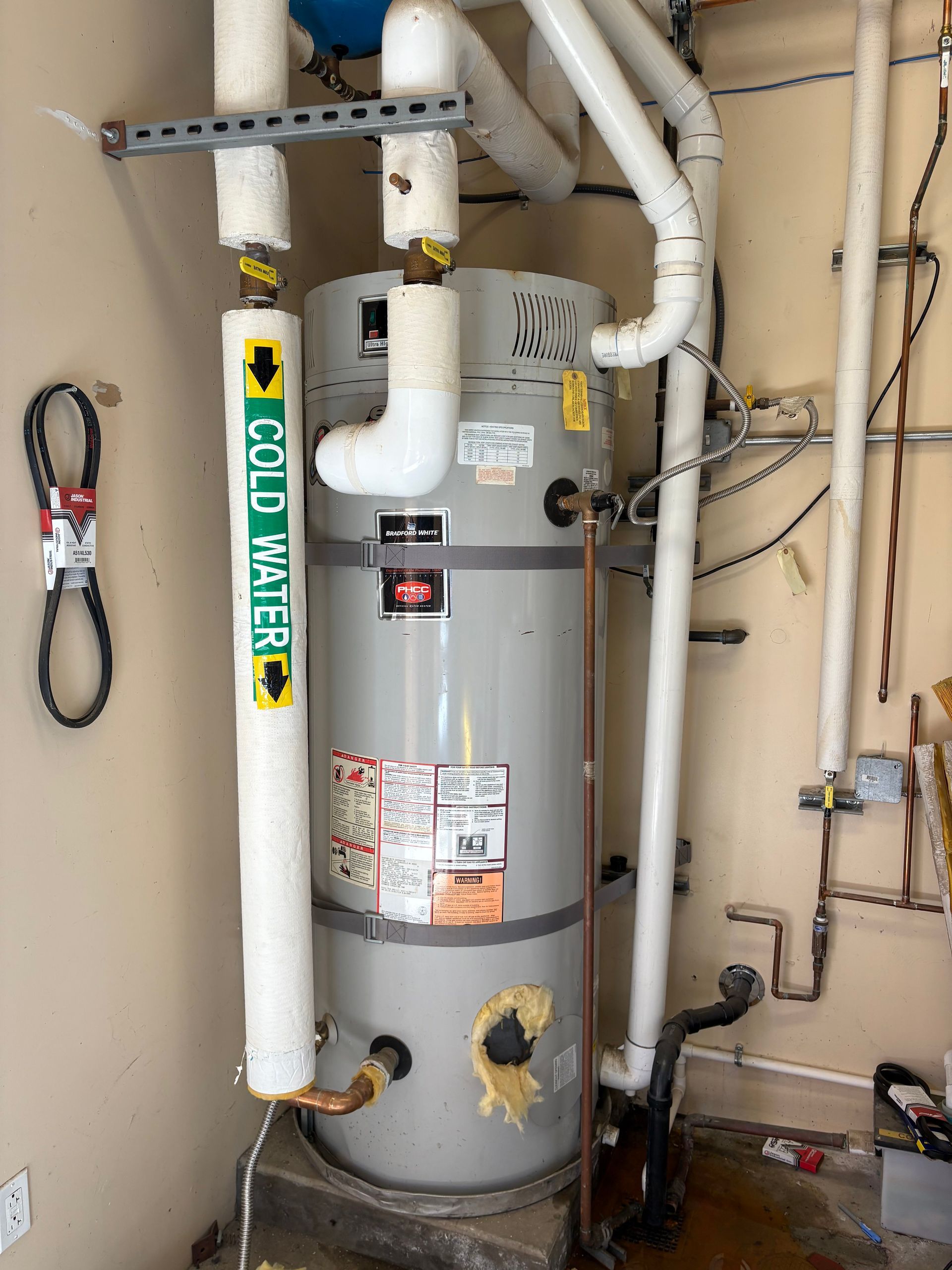 A gray residential water heater tank stands in a utility room, wrapped in insulated white pipes and labeled cold water.