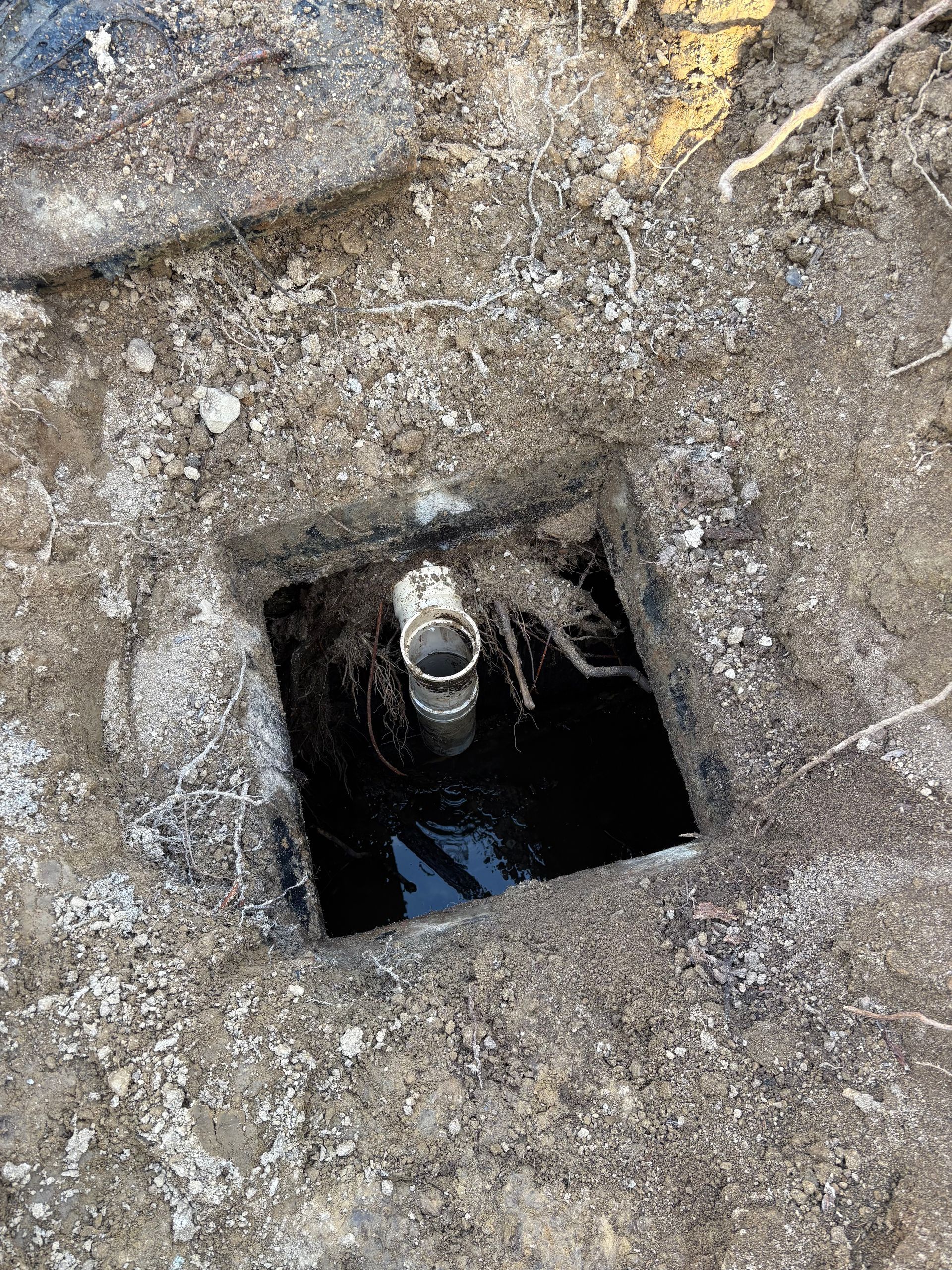 A vertical pipe sits inside a square, dirt-filled hole with standing water at the bottom.