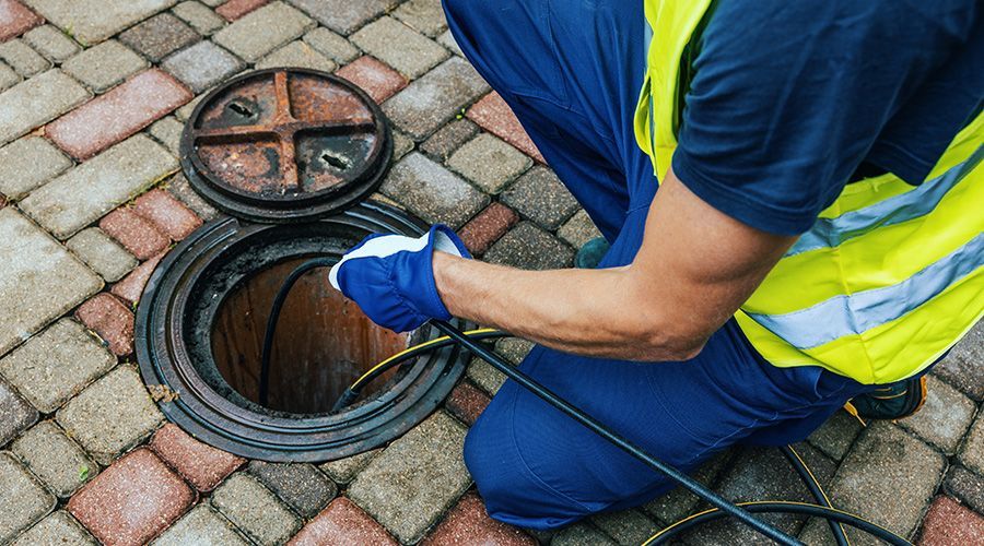 A worker in a yellow safety vest feeds a drain cleaning cable into an open sewer manhole on a brick-paved surface.