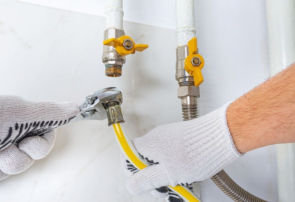 A person wearing gloves uses a wrench to tighten a yellow hose onto a wall-mounted valve in a white tiled area.