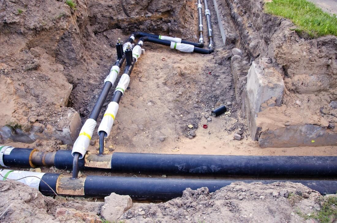 Black pipes with insulated joints lie in an open dirt trench during utility or construction work.