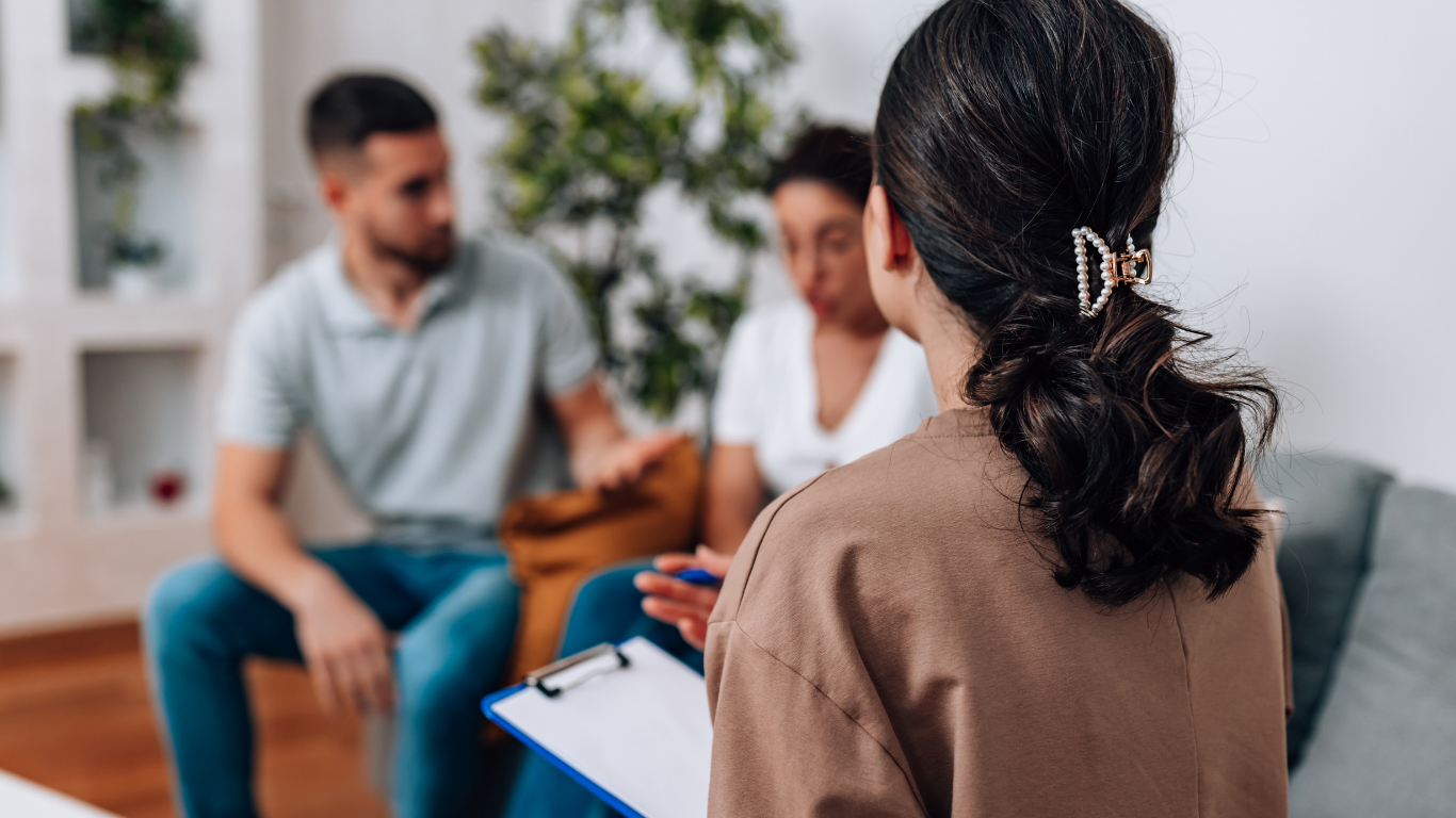 Therapist with clipboard consults with a blurred couple on a sofa; indoors, neutral colors.