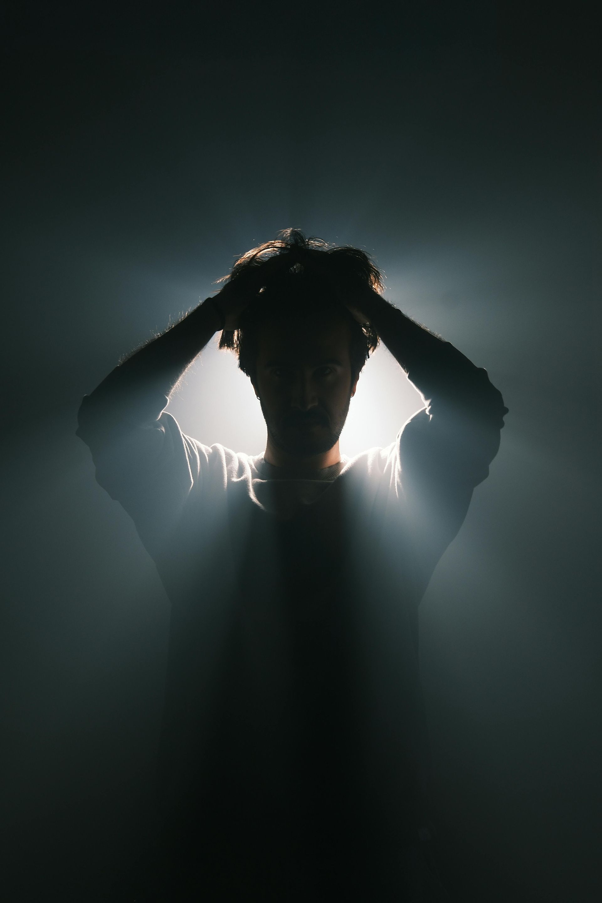 Silhouetted person with hands on head, backlit by bright light in a dark, smoky setting