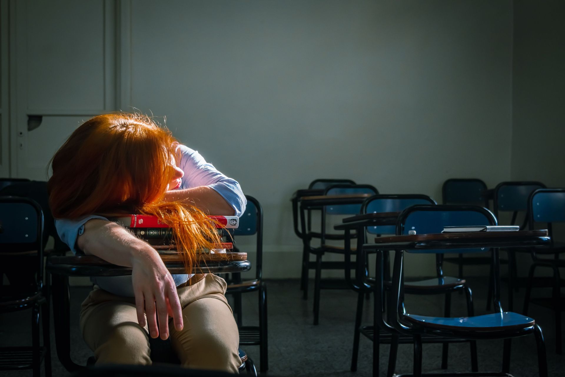 Person with red hair rests at a desk with empty blue chairs showing signs of emotional exhaustion