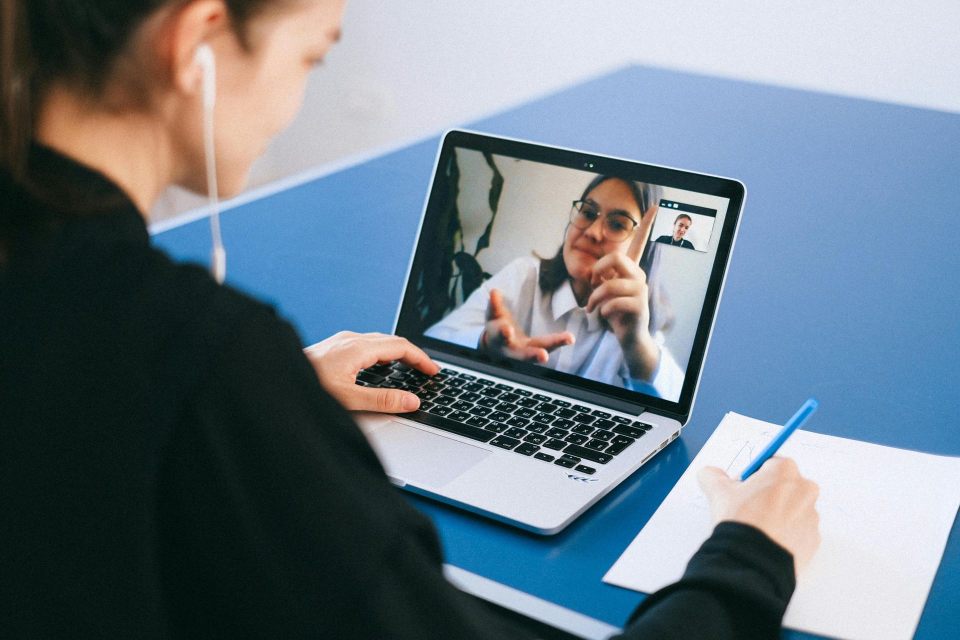 Woman on laptop video call, taking notes on paper, blue table.