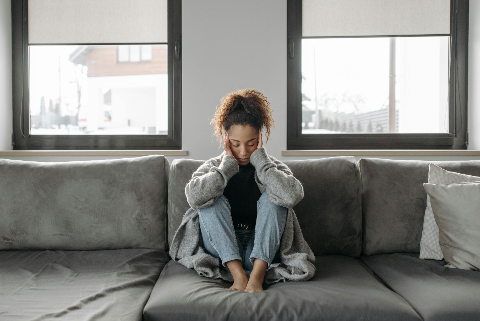 Person sitting on a gray couch with head in hands in a bright living room