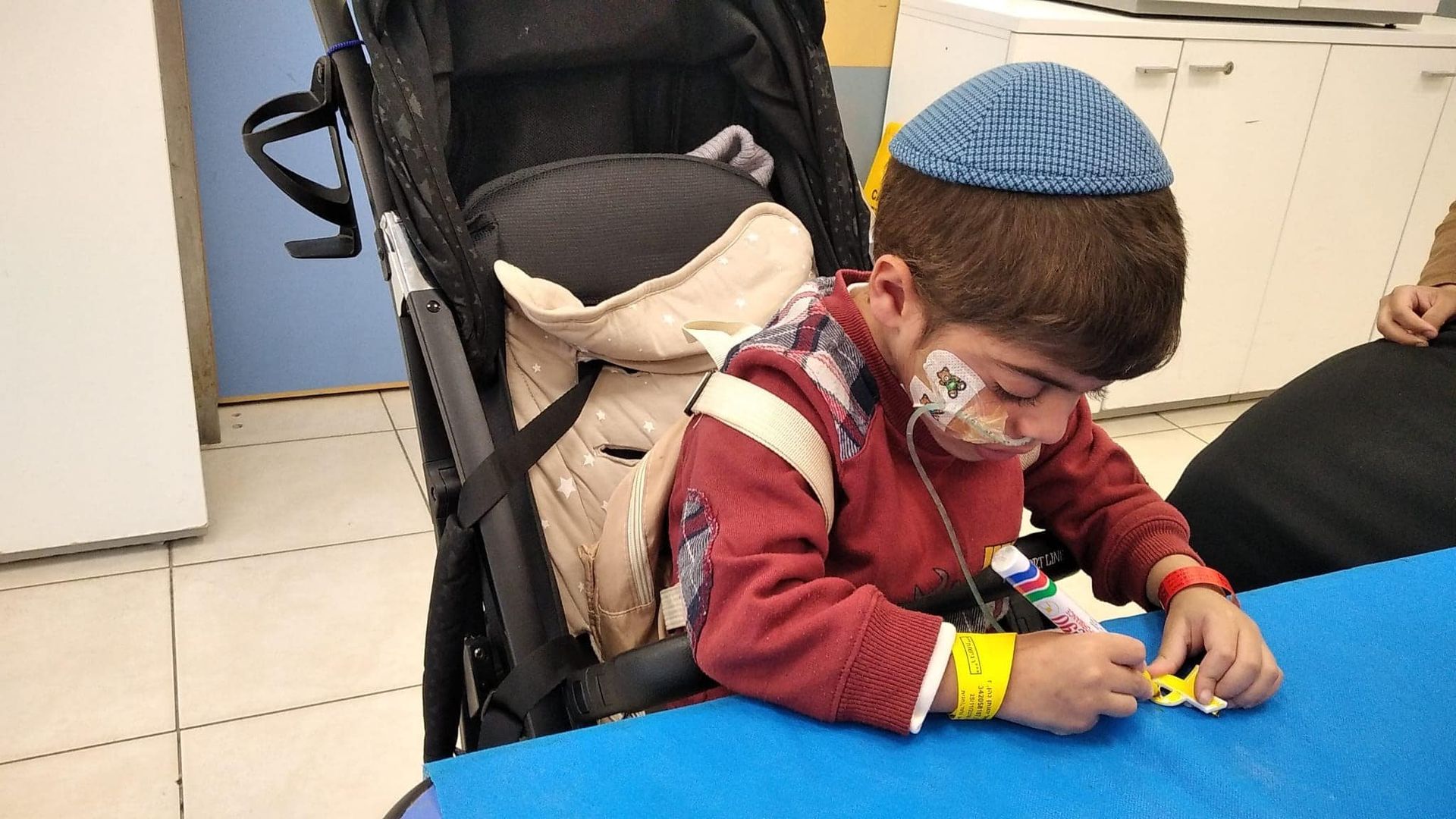 A young boy wearing a kippah is sitting at a table with a stroller in the background.