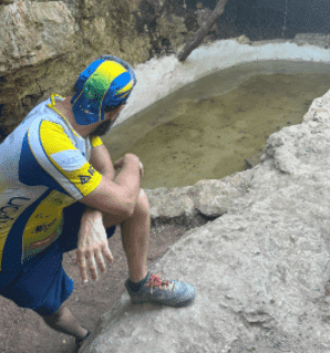 A man wearing a blue and yellow helmet sits on a rock near a body of water.