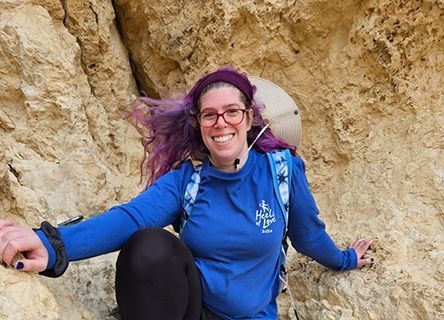 A woman with purple hair and glasses is sitting on a rock.