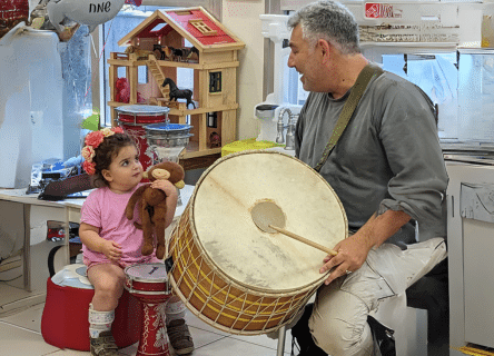 A man is playing a drum while a little girl holds a teddy bear.