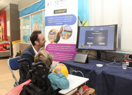 A man is sitting next to a child in a wheelchair in front of a computer screen.