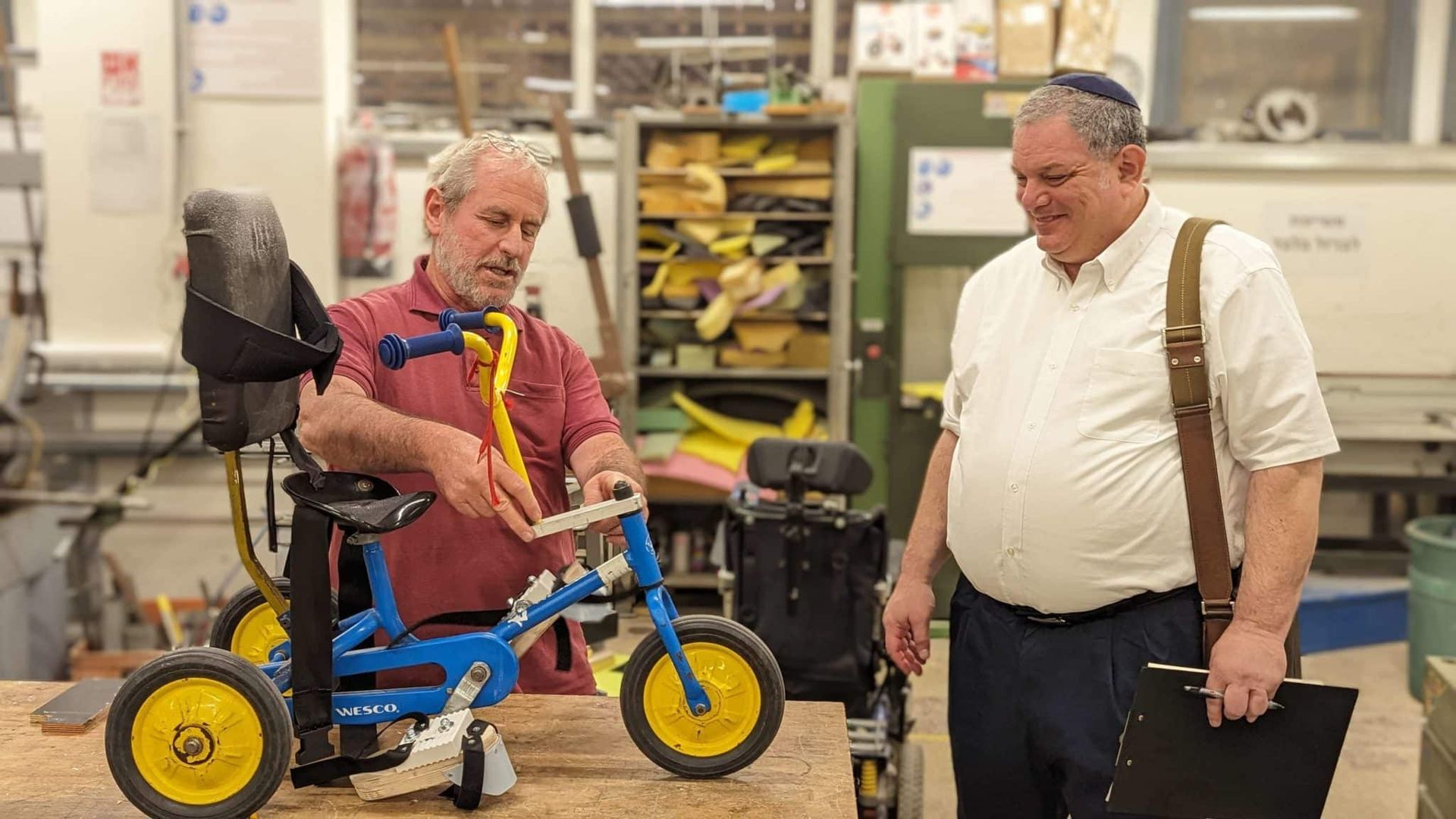Two men are standing next to each other in a workshop looking at a tricycle.