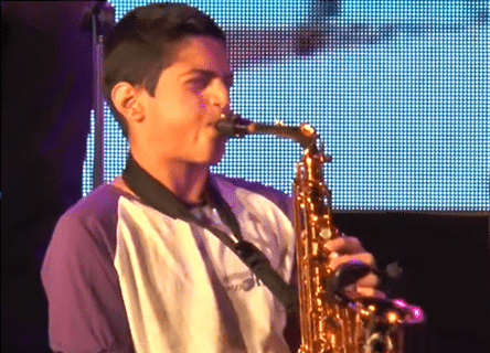 A young man is playing a saxophone in front of a large screen
