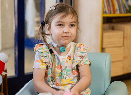 A little girl is sitting in a chair with a necklace around her neck.