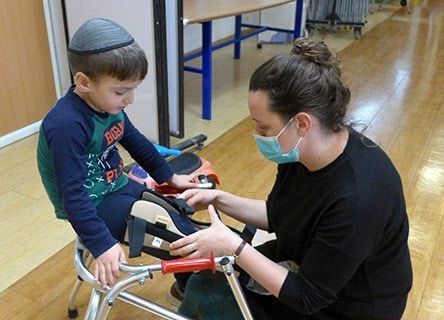 A woman wearing a mask is helping a young boy with a walker.