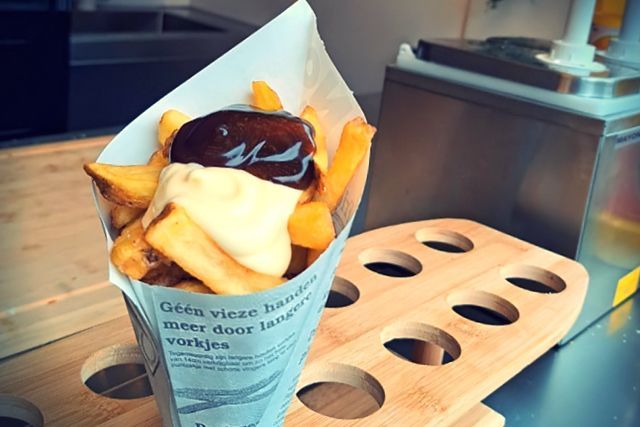 Een papieren puntzak gevuld met frietjes, bedekt met mayonaise en kerriesaus, in een houten houder.