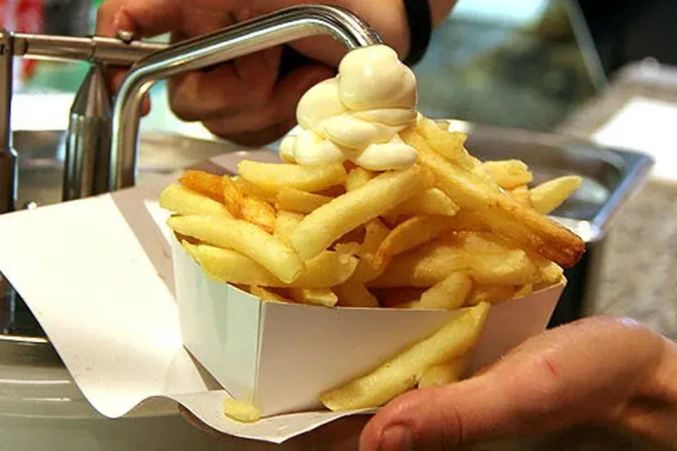 Een hand houdt een wit papieren bakje vast met goudbruine frietjes, bedekt met een flinke klodder witte mayonaise.