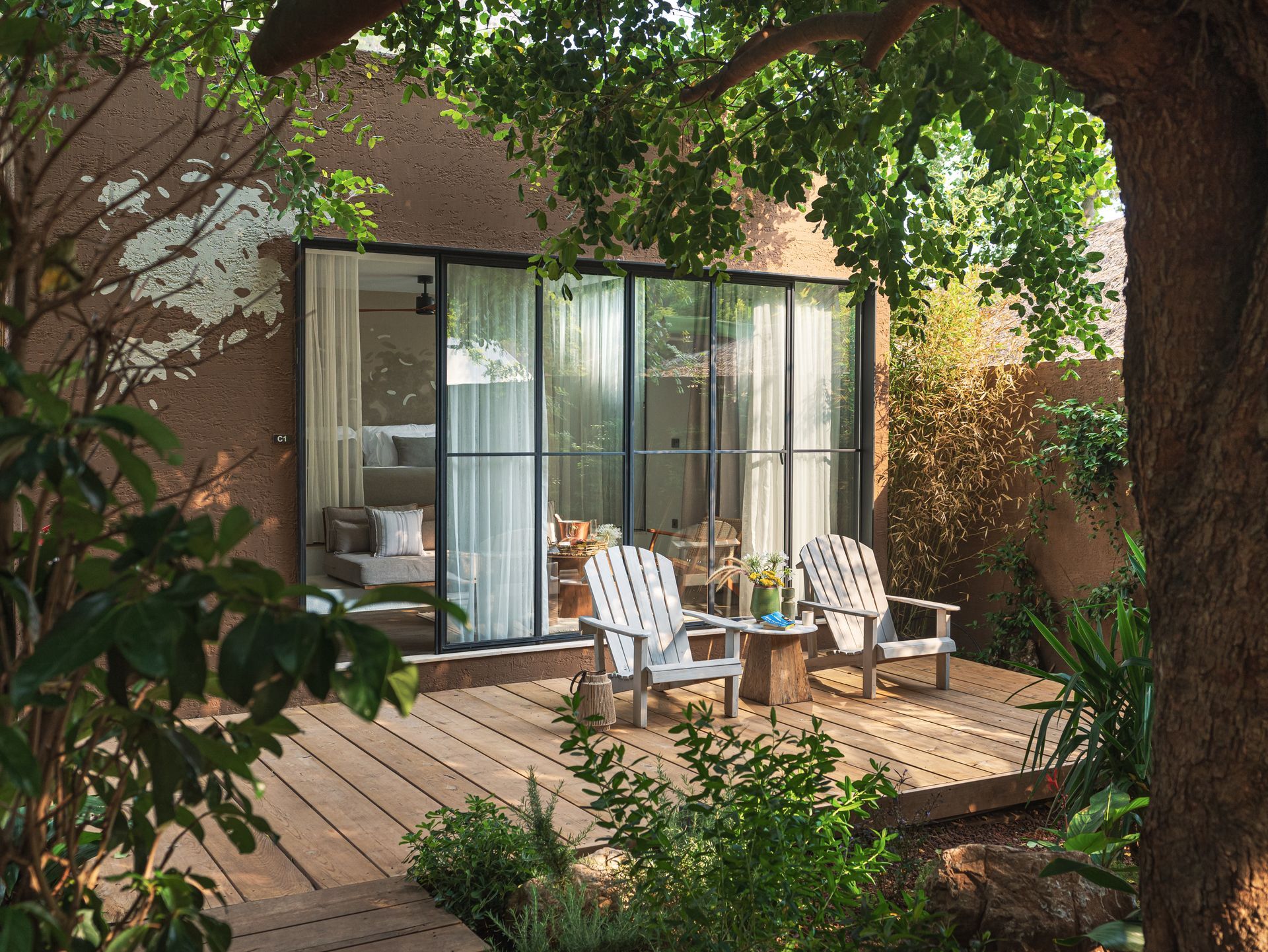 A luxury resort patio with two lounge chairs, decorative pillows, and a small pool in front of a bedroom with a lush view.