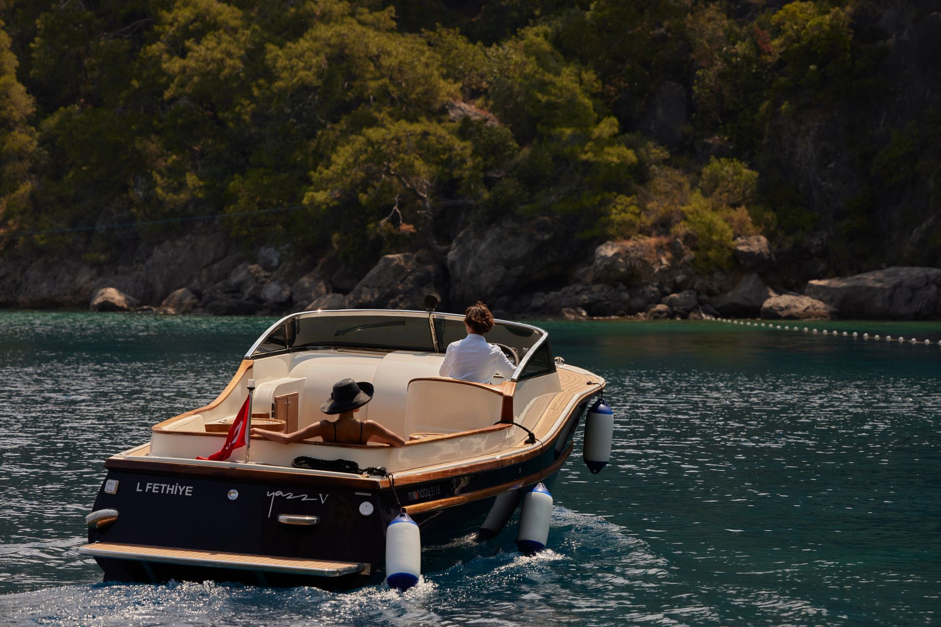 A dark-hulled motorboat with two people on board cruises on clear blue water near a rocky, forested shoreline.