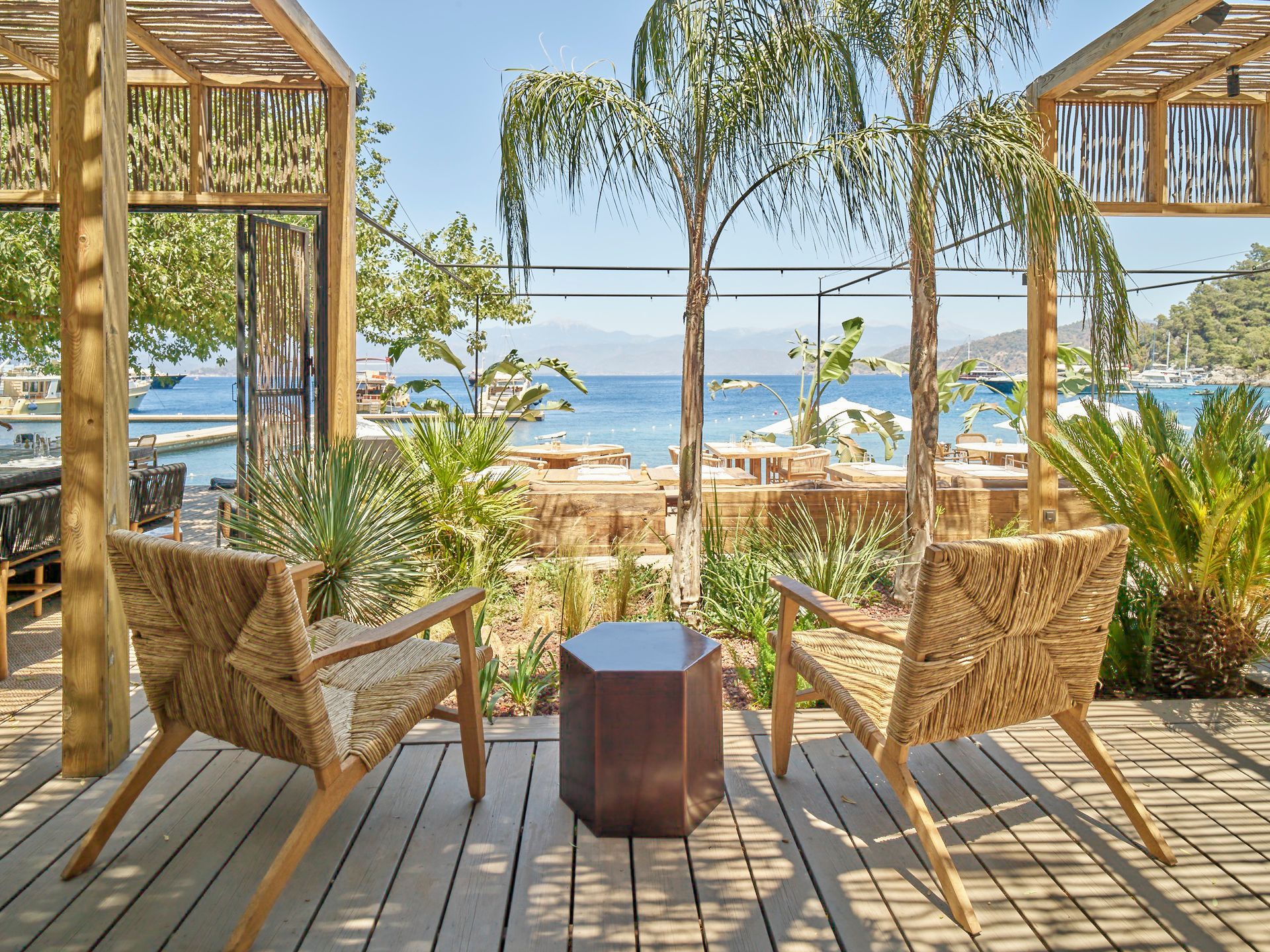 Two woven chairs face a calm blue sea, separated by a small brown side table on a wooden deck under palm trees.