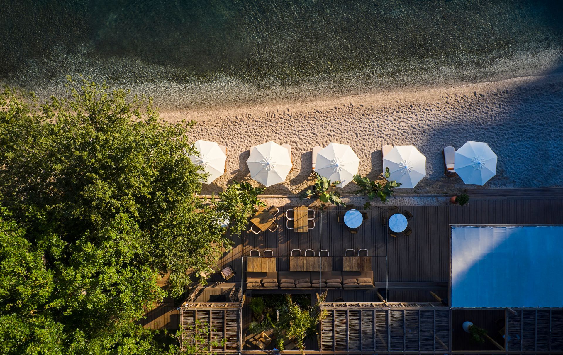 Aerial view of a beach with white umbrellas, outdoor furniture on a deck, and trees along the shore.