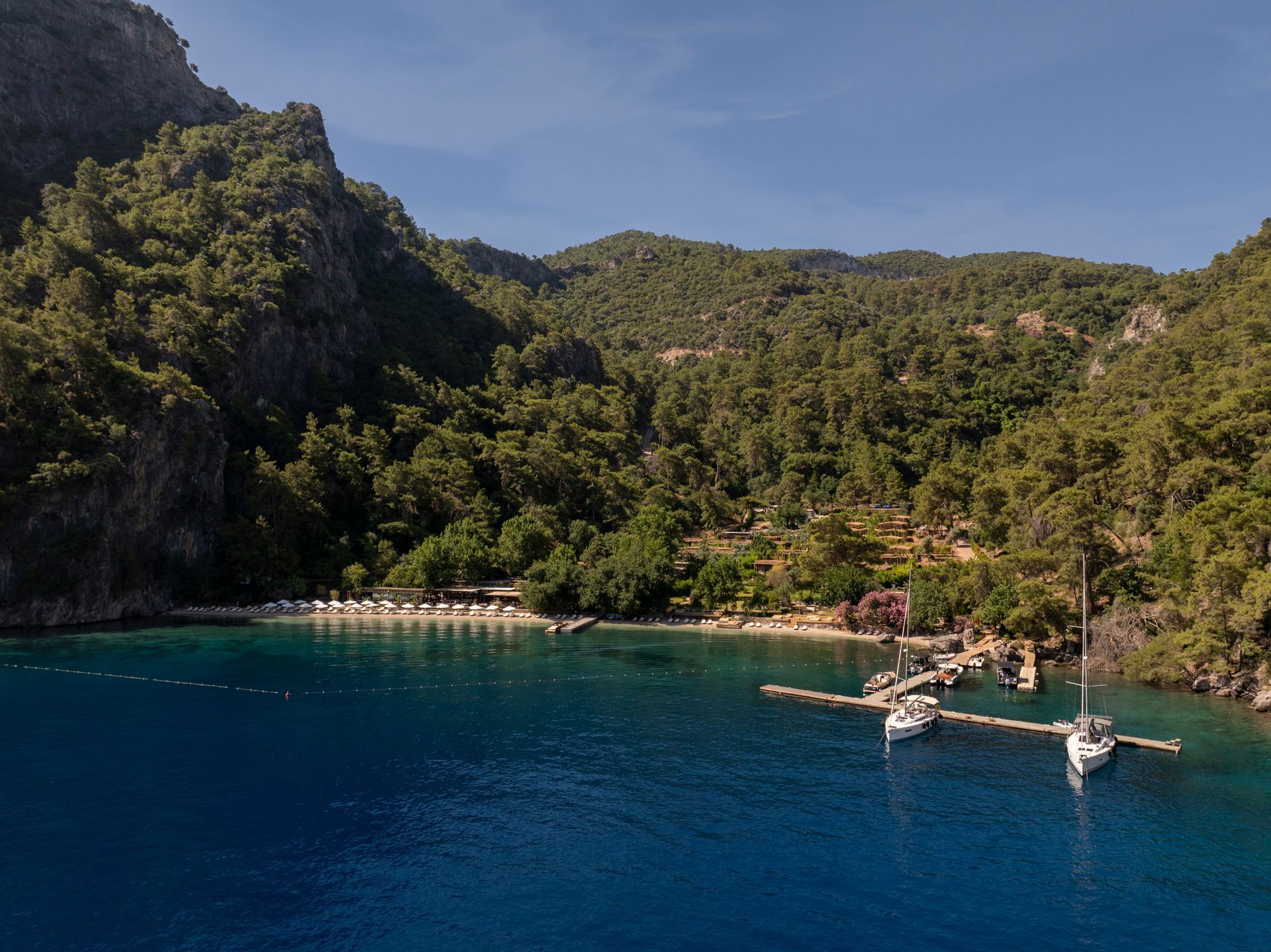 An aerial view of a secluded blue bay surrounded by forested mountains, with sailboats docked at a pier near a beach.