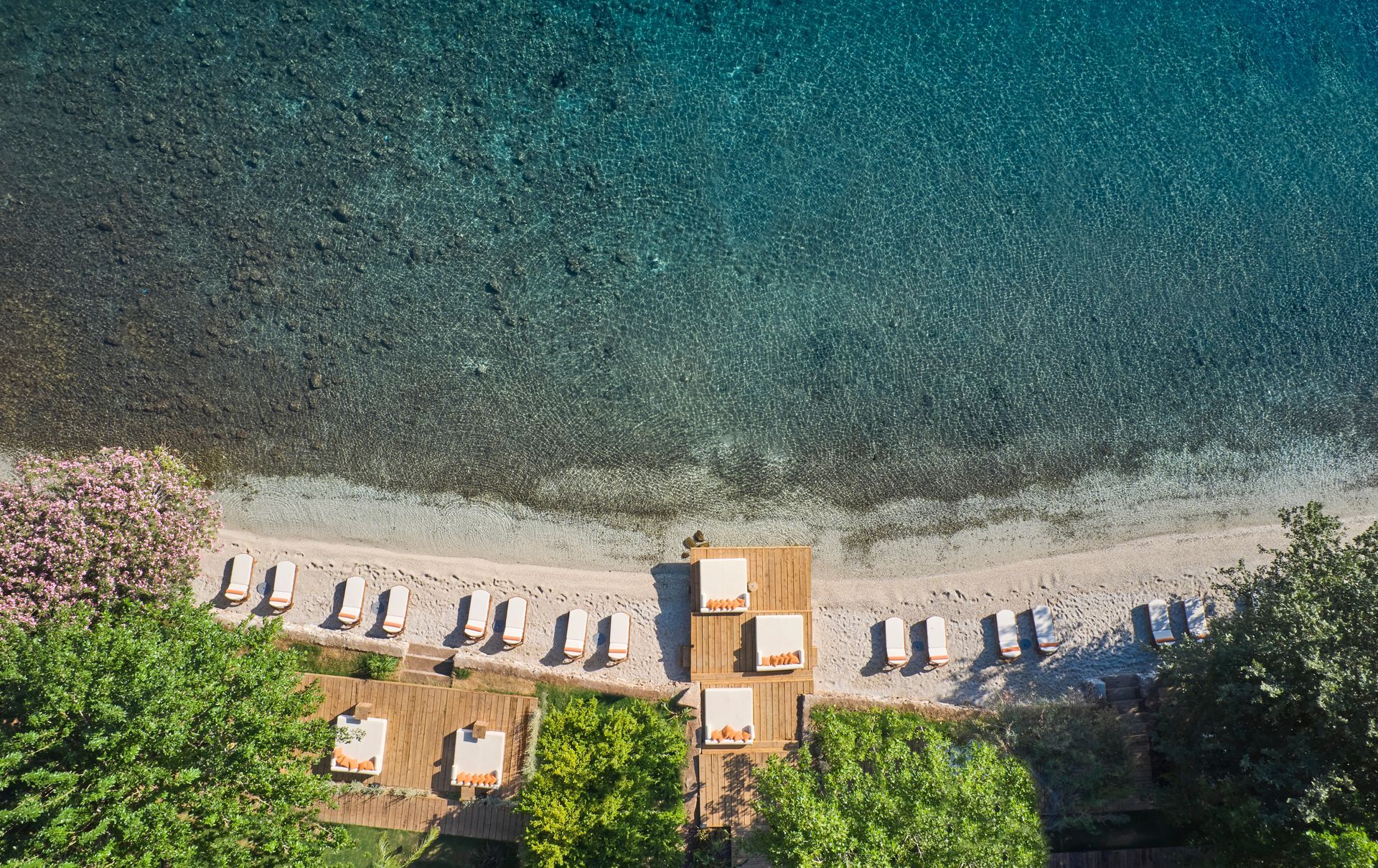 Aerial view of a sunny beach with lounge chairs, a wooden deck, and lush trees lining the turquoise shoreline.