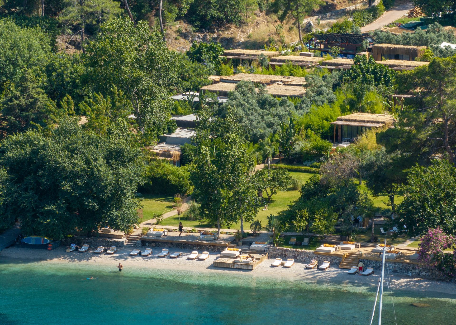 Aerial view of a coastal resort with terraced buildings, lush green trees, and lounge chairs on a pebble beach.