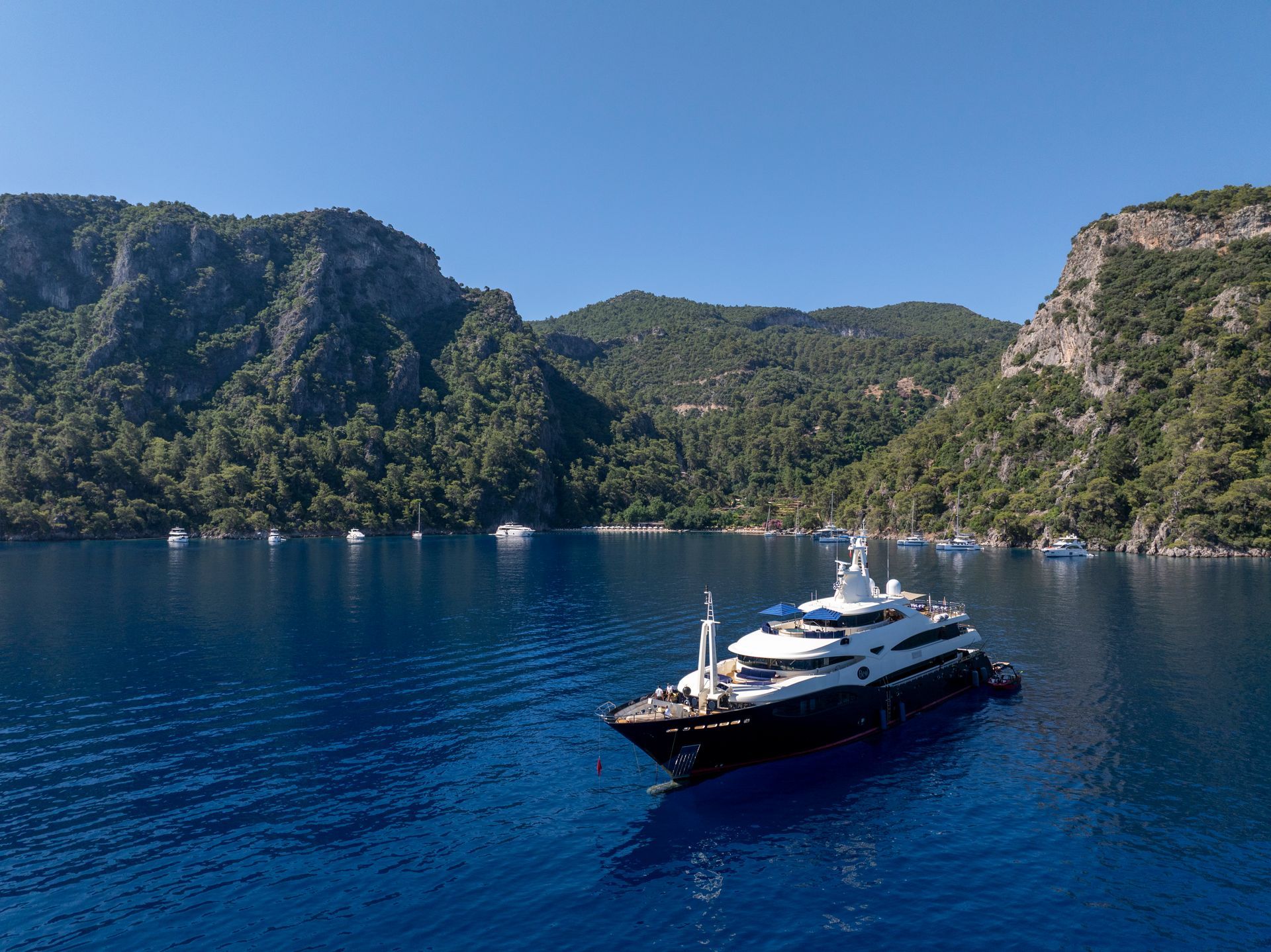 A large, modern motor yacht cruises on deep blue water in a cove surrounded by steep, tree-covered hills.
