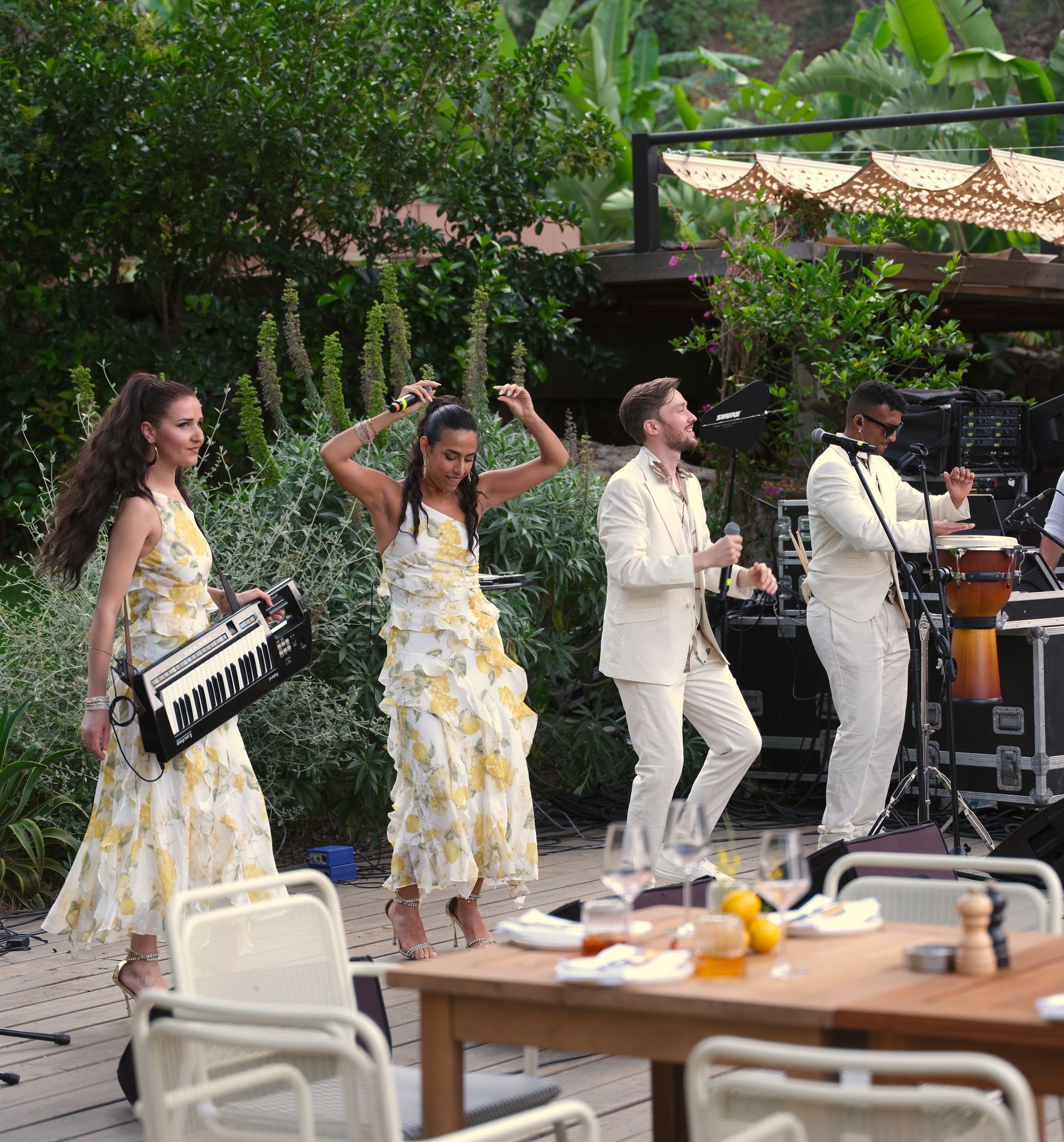 A band plays music outdoors, with two people in floral dresses and two in white suits performing on a patio.