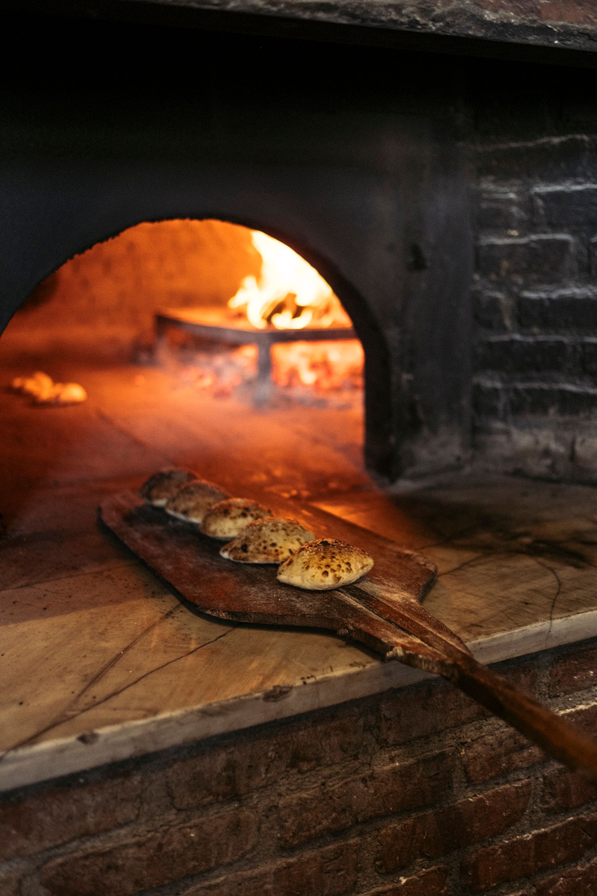 A wooden peel holds several round, seasoned flatbreads in front of a rustic, wood-fired brick oven with glowing flames.