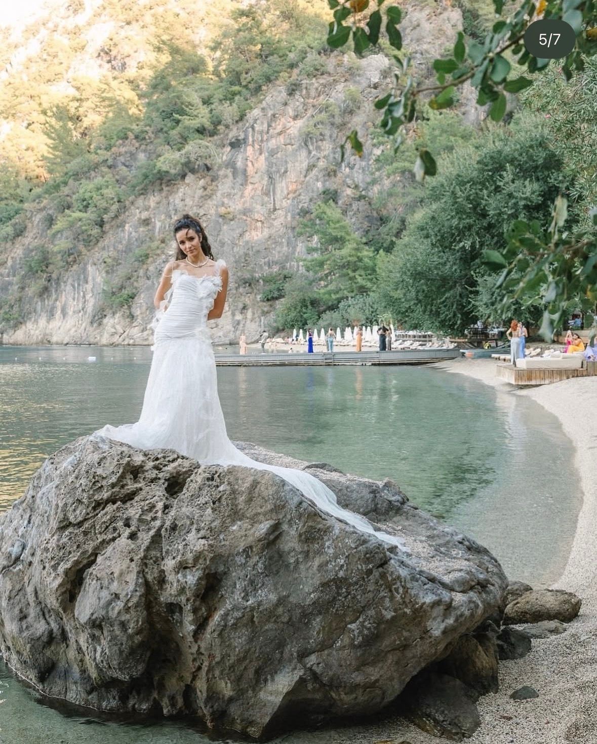 A person in a white wedding gown stands on a large rock at the edge of a serene bay with cliffs and trees in the background.