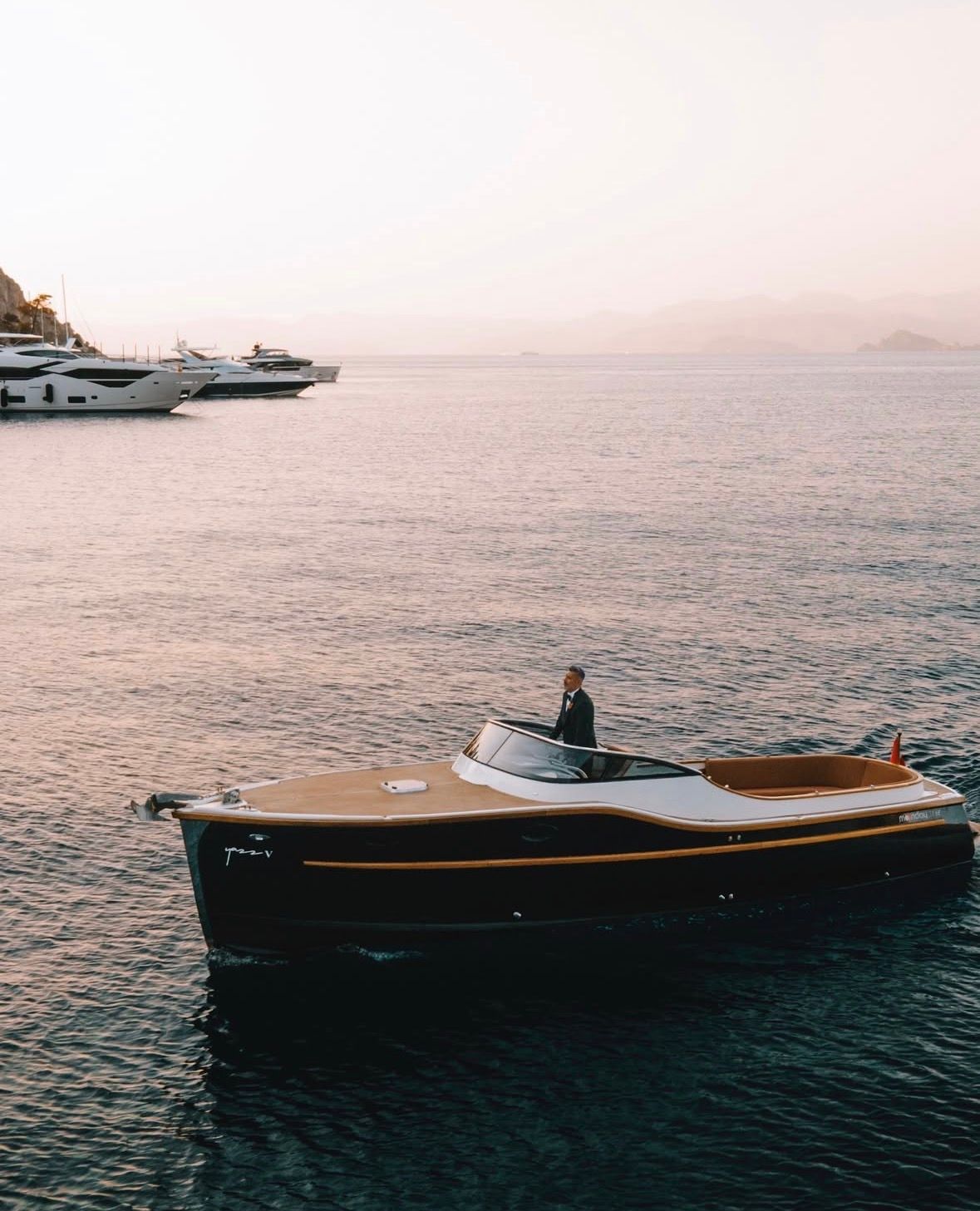 A small, classic wooden motorboat with a dark hull cruises on calm water at sunset, with larger yachts in the background.