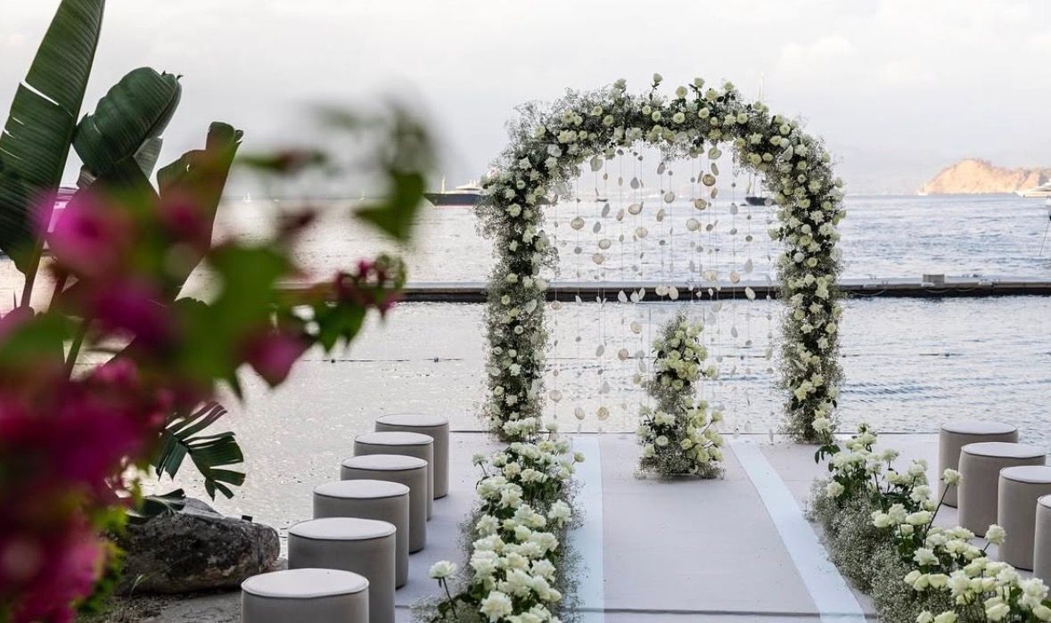 A wedding ceremony setup with a white floral arch, a central aisle, and grey cushioned stools by a calm body of water.