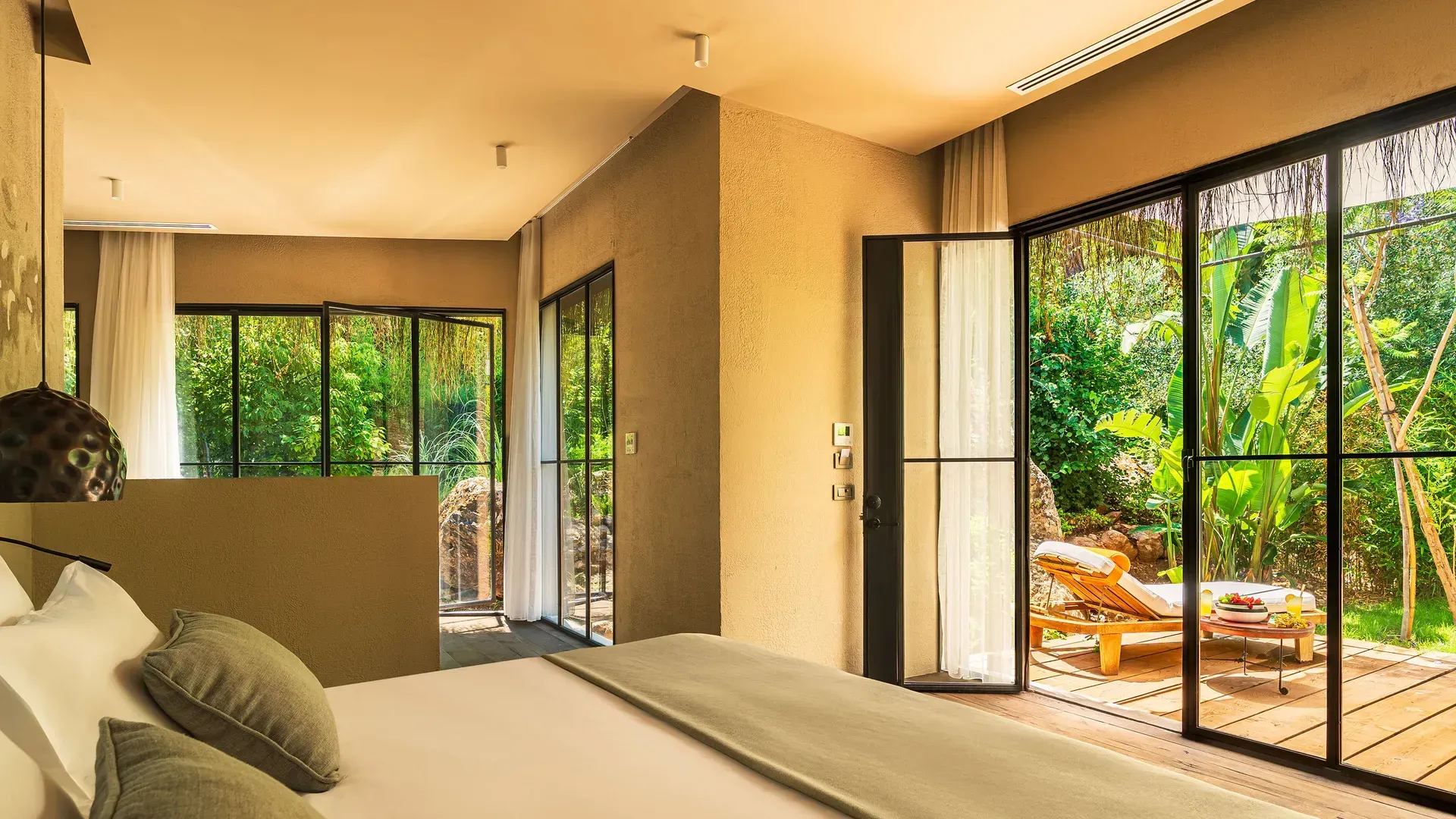 Modern bedroom with large glass doors opening to a lush garden, wooden deck, and a lounge chair, bathed in warm light.