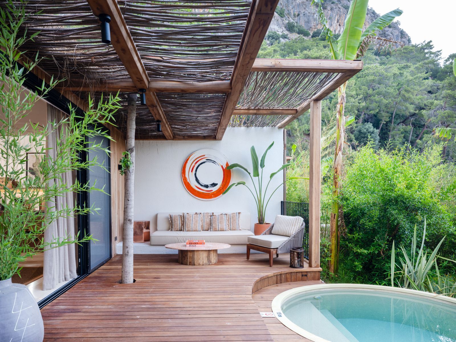 A patio with wood decking, a circular pool, seating, and a pergola, set against a lush, mountainous backdrop.