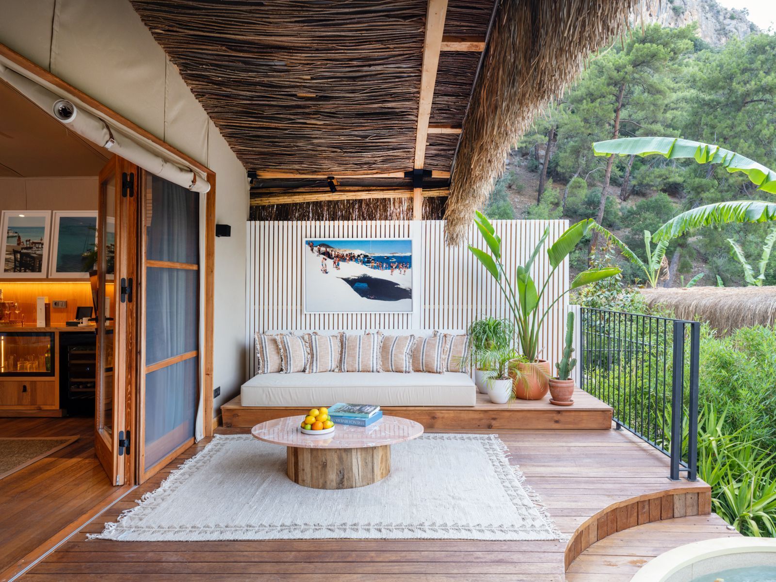 An outdoor wooden deck with a white sofa, a round pink coffee table, and lush tropical plants overlooking a forest.