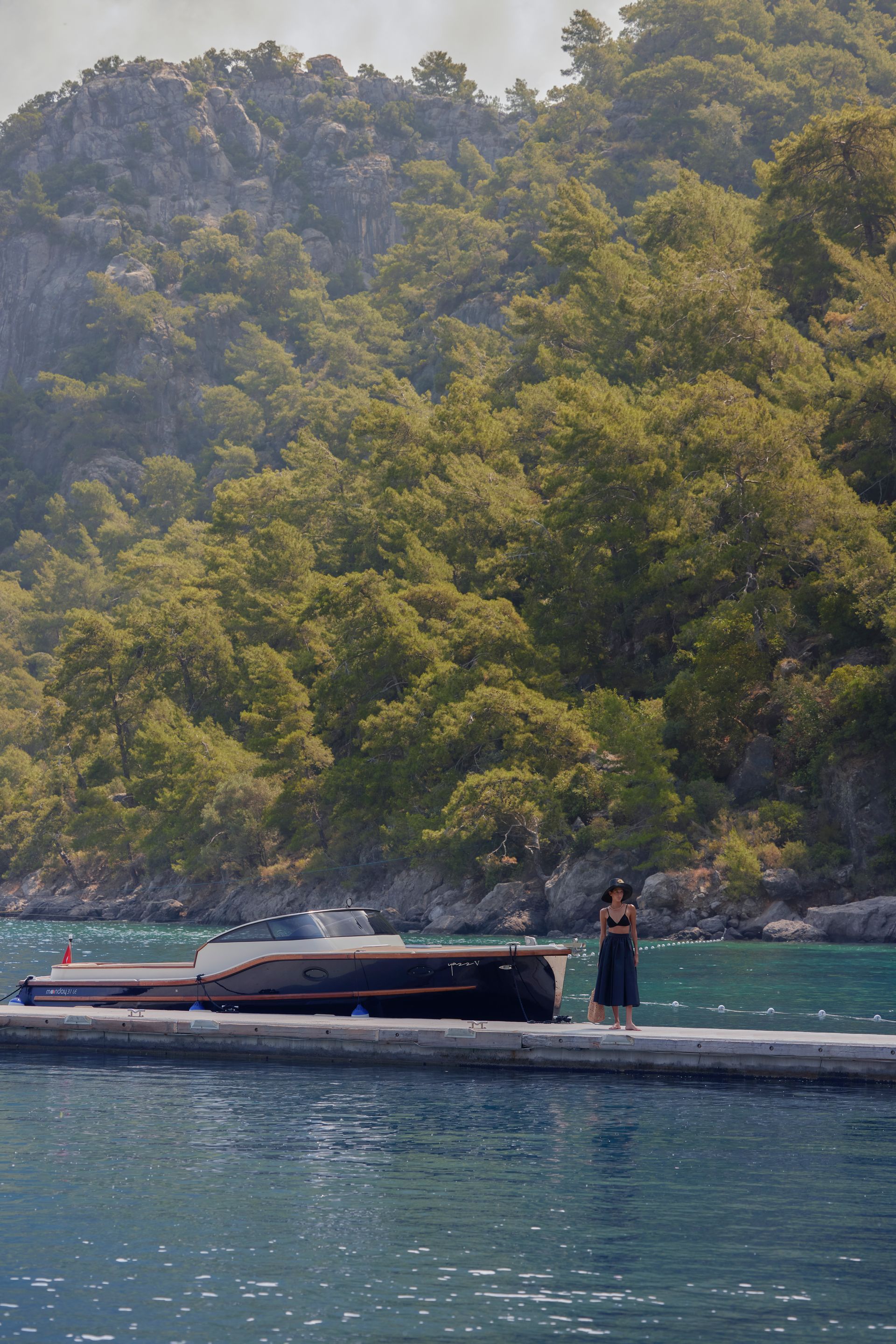 A person stands on a stone pier next to a dark-hulled motorboat, with a forested mountain rising in the background.