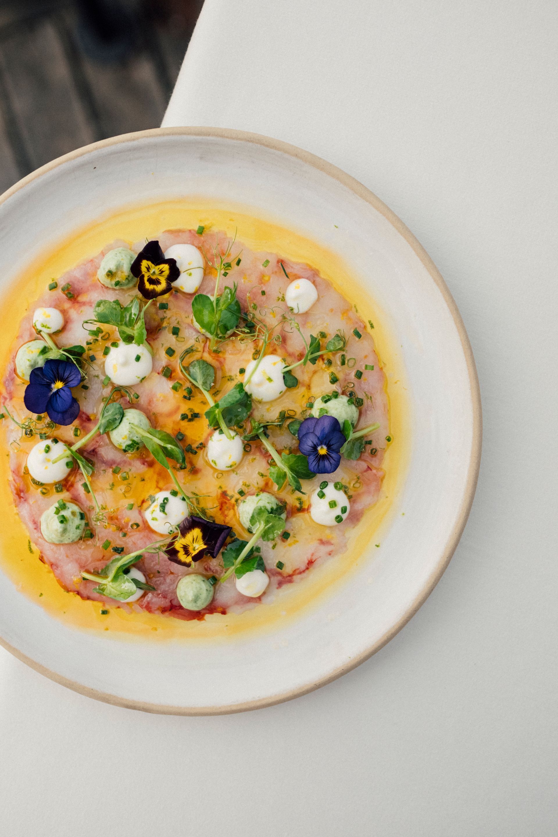 A top-down view of a circular raw fish carpaccio topped with dollops of cream, fresh herbs, and edible purple flowers.